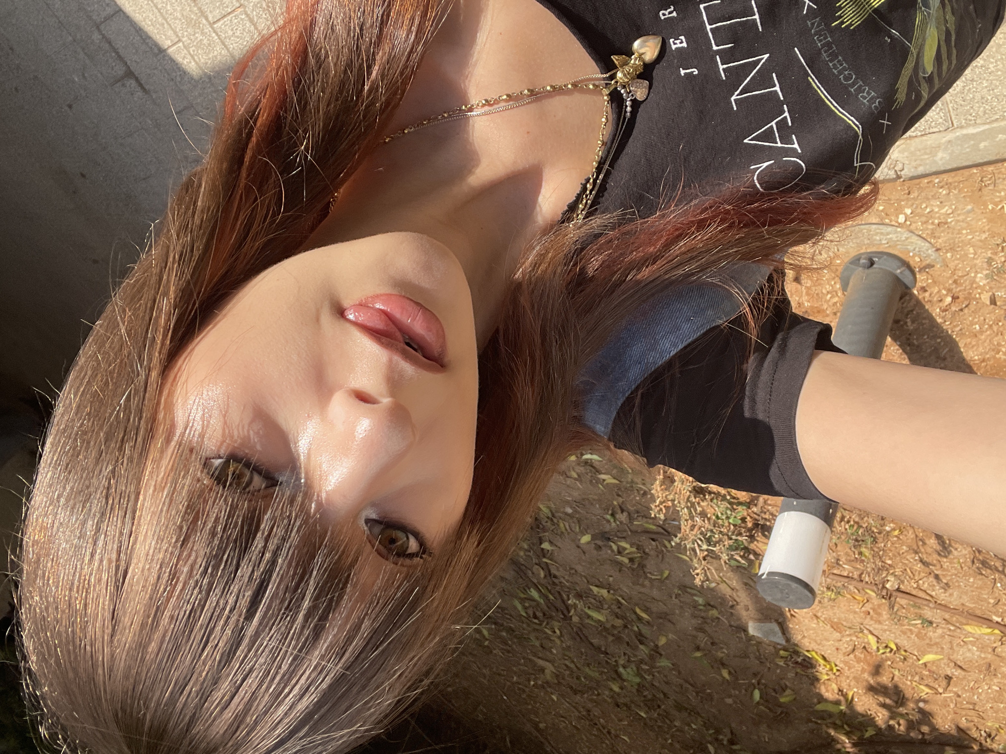 A young woman with long, layered brown hair with auburn highlights is taking a selfie outdoors in Raanana, Israel. The sunlight catches her hair and face, creating a glow. She is wearing a black t-shirt with white text that reads "JER CANTIL" and a yellow graphic. She is also wearing a denim jacket and black shorts. She has on multiple necklaces, one with a heart and angel charm. Her makeup includes eyeliner and lip gloss. In the background, there is dirt, dry leaves, and a paved walkway. A metal pole with a white and gray cylindrical object on top is visible in the foreground.