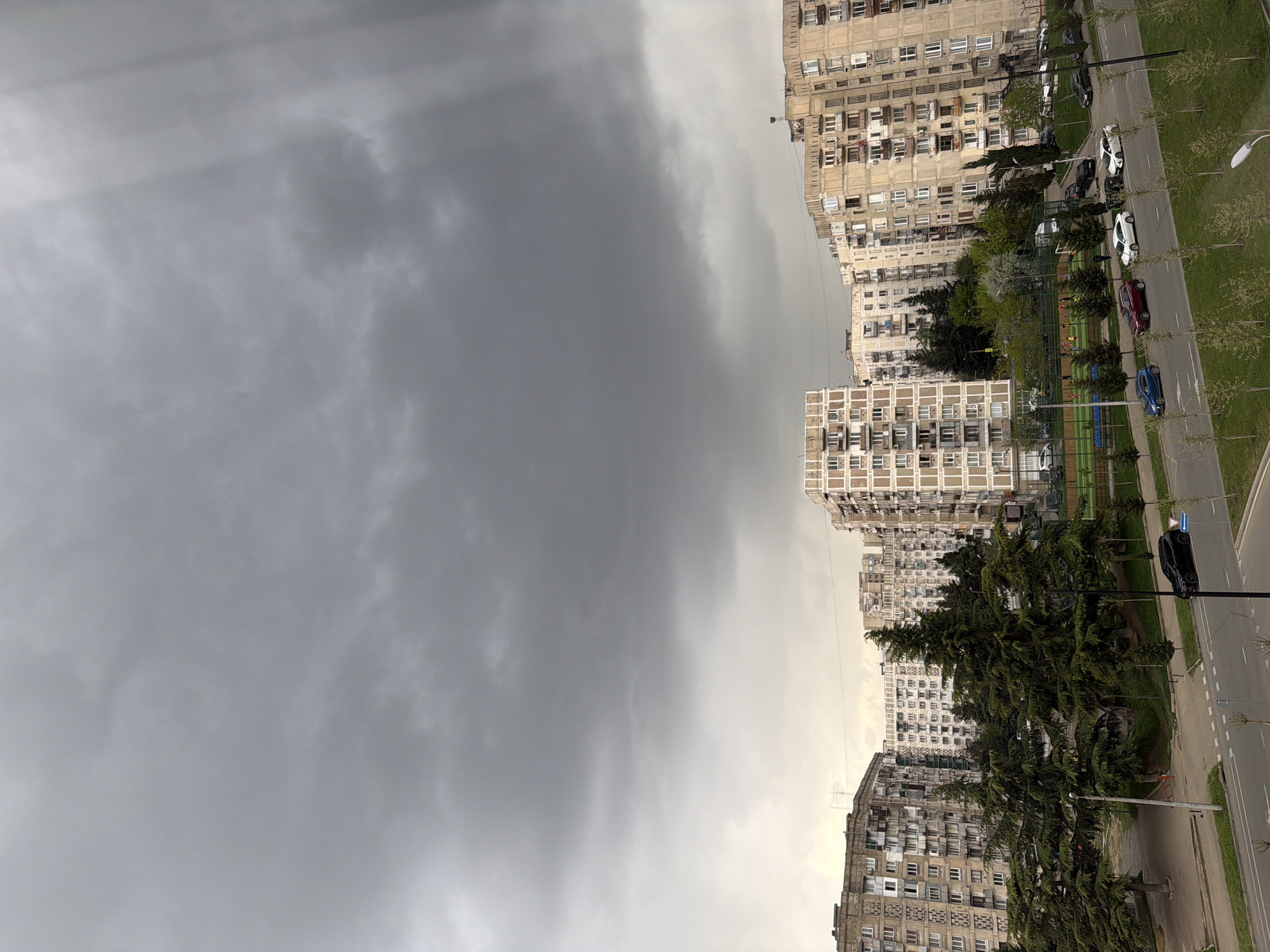 The image shows a view in Tbilisi, Georgia, characterized by Soviet-era apartment blocks and a cloudy, overcast sky. The buildings are high-rise with repetitive patterns of windows. A street runs along the side, with parked cars and trees lining the road. A children's playground is visible between the buildings. The sky is filled with heavy, dark clouds, indicating possible rain. A street lamp is visible in the foreground.