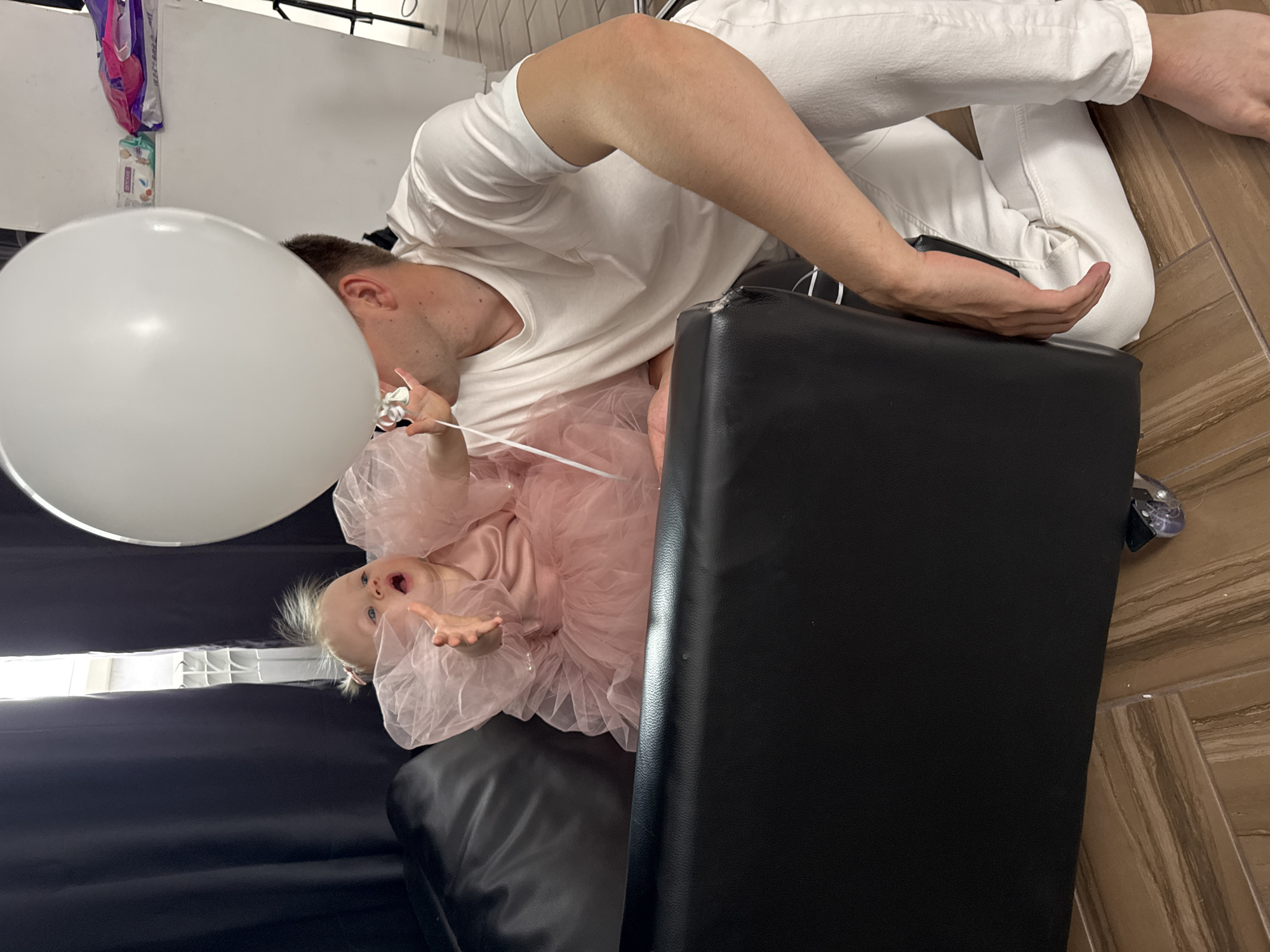 The image shows an adult and a young child. The adult is wearing white jeans and a white t-shirt. The child is wearing a light pink ruffled garment and is holding a large white balloon. The adult and child are positioned near a black leather chair or ottoman. The background includes what appears to be a white wall or panel. The floor has a wood-like pattern. Based on the prompt information, the location is in the city of Chaikovsky, Russia.