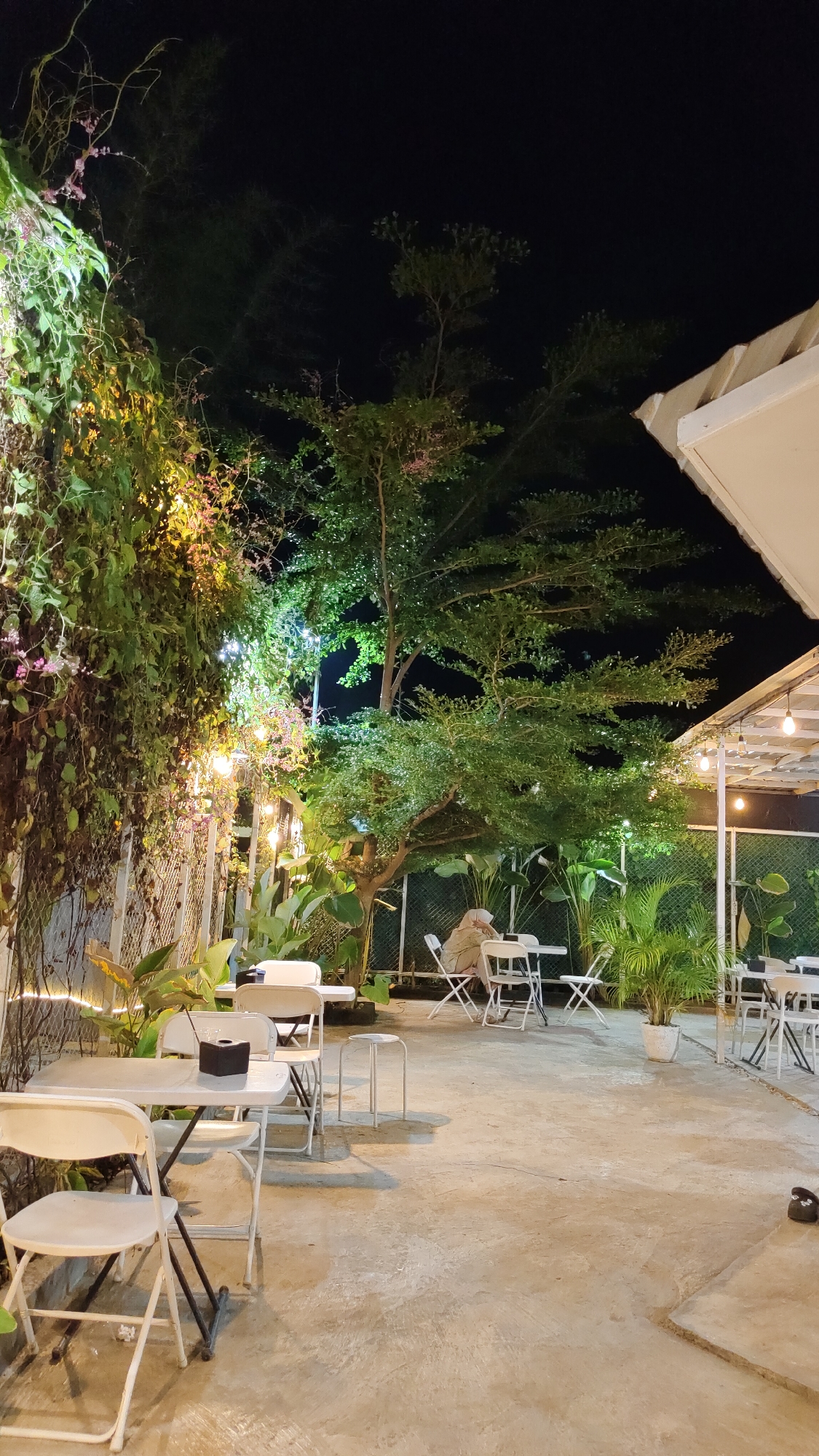This is an outdoor nighttime scene at a cafe in Keudebayu, Indonesia. The foreground features several white folding chairs and tables, some with black napkin holders. A person wearing a hijab is seated at a table in the middle ground, partially obscured by foliage. The background is dominated by lush green trees and dense vegetation, illuminated by string lights. The overall atmosphere is tranquil and inviting, typical of a relaxing evening at a cafe.