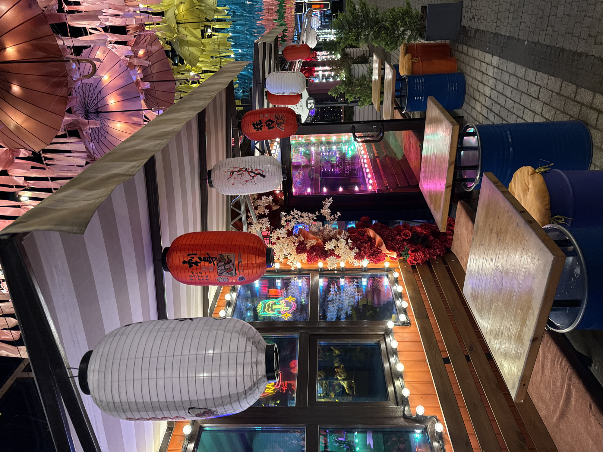 This image depicts an outdoor seating area of a restaurant or cafe, likely in Krasnodar, Russia, given the context. The scene is illuminated by artificial lights and decorated with numerous hanging lanterns and paper umbrellas in various colors. The seating consists of wooden tables with wooden barrel-style bases, some of which are painted blue and orange. Cushions, in an orange hue, are placed on top of the barrel bases to serve as seats. In the background, a wooden bench with red cushions runs along a wall featuring a large window with orange lighting. Decorative floral arrangements, predominantly red roses and white flowers, are visible. The ground is paved with grey cobblestones.