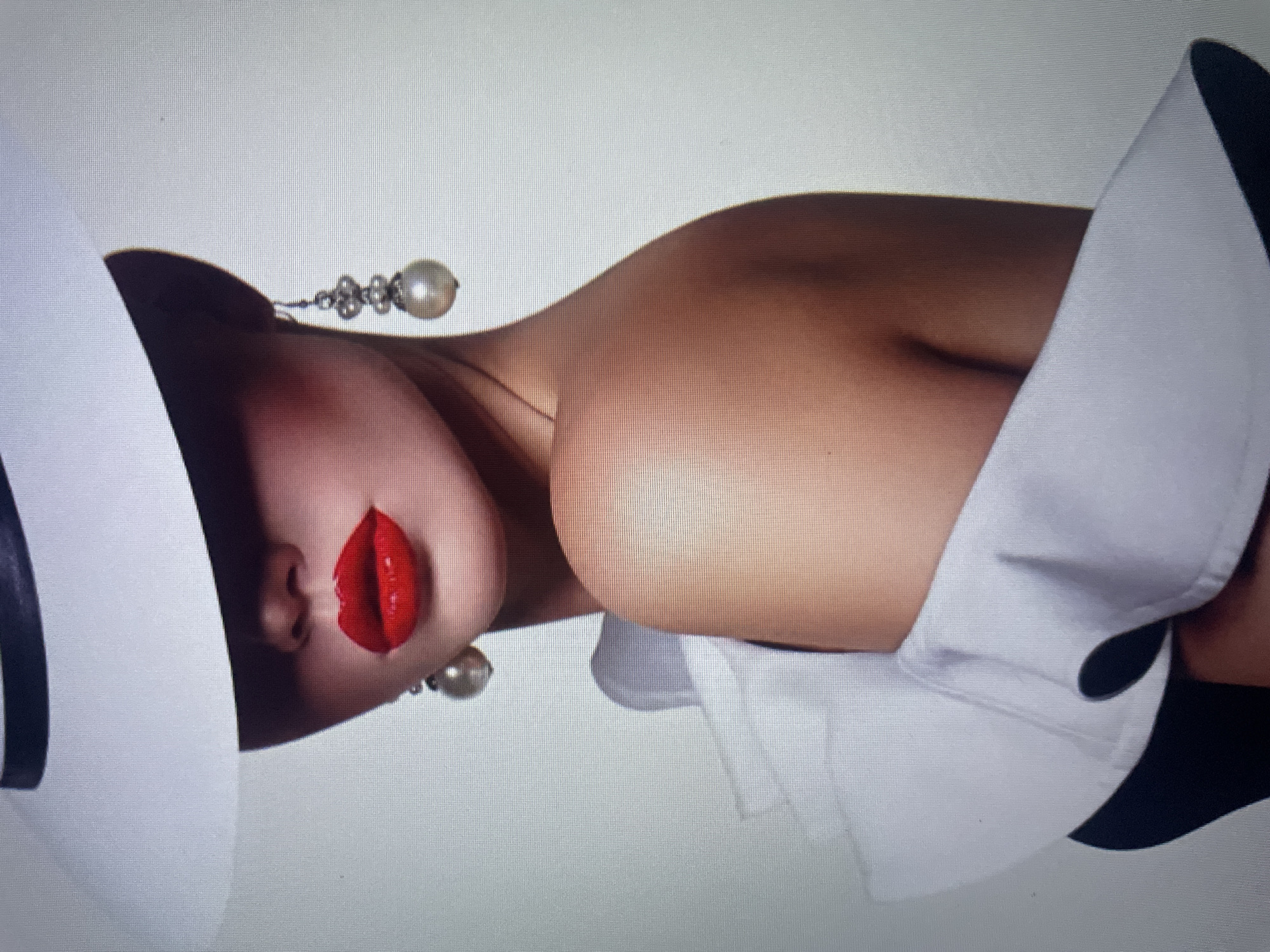 The image is a medium shot of a woman's head and shoulders, taken against a plain, light gray background. She is wearing a wide-brimmed white hat that casts a shadow over her eyes and the top of her face. Her lips are painted a vibrant red, and she is wearing a pair of ornate, dangling earrings, each featuring a large pearl at the bottom. The woman's shoulder is bare, and she is wearing a white, ruffled garment, possibly a dress or a blouse, with a black garment visible underneath. The lighting is soft and highlights the contours of her face and shoulder. There is no identifiable location within the city of Kołobrzeg, Poland, or any other specific city.