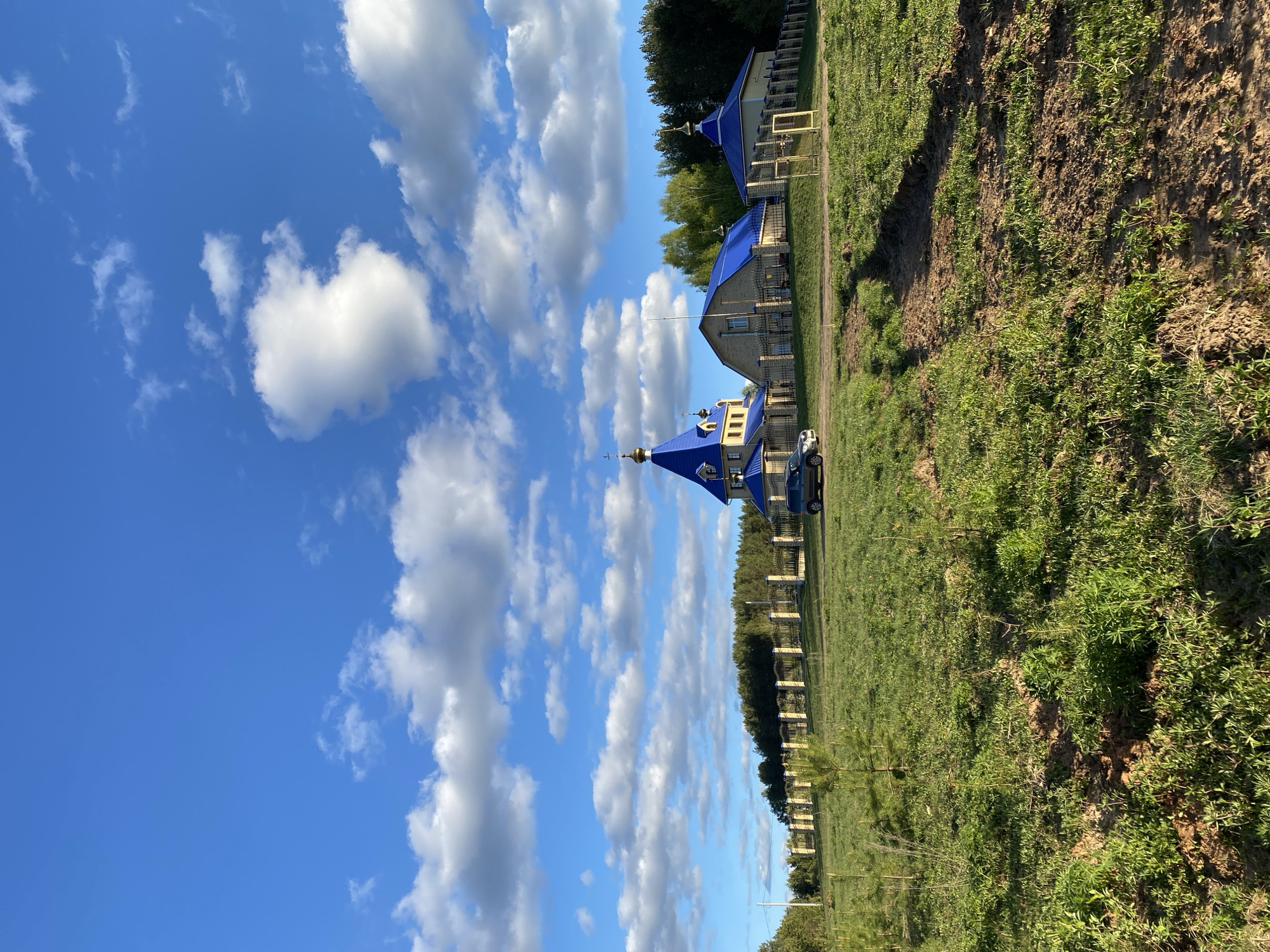 The image captures a landscape featuring a series of structures with distinctive blue roofs, likely churches or religious buildings, situated along a line parallel to a fenced perimeter. The structures are located in Chaikovsky, Russia. The environment consists of grassy areas and patches of cultivated soil, possibly recently plowed or worked. A clear blue sky is filled with numerous white clouds, creating a visually striking backdrop. A vehicle is seen parked near one of the buildings. A fenced perimeter encloses the buildings. The scene suggests a serene or rural setting within the urban region of Chaikovsky.