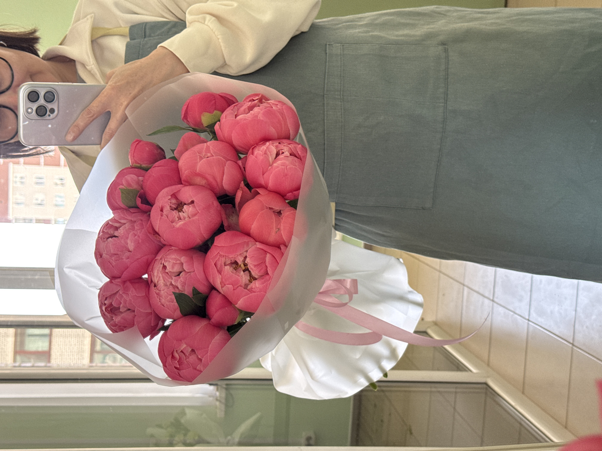 A person is taking a selfie with a bouquet of pink flowers in front of a window in what appears to be an indoor location, likely a room. The person is wearing glasses, a light-colored shirt, and a green overall dress. The smartphone held by the person has multiple camera lenses visible. The bouquet is wrapped in white paper with a pink ribbon. The background shows a building through the window. Based on the architectural style of the buildings visible through the window and the urban setting, the location is possibly in the city of Izhevsk, Russia.