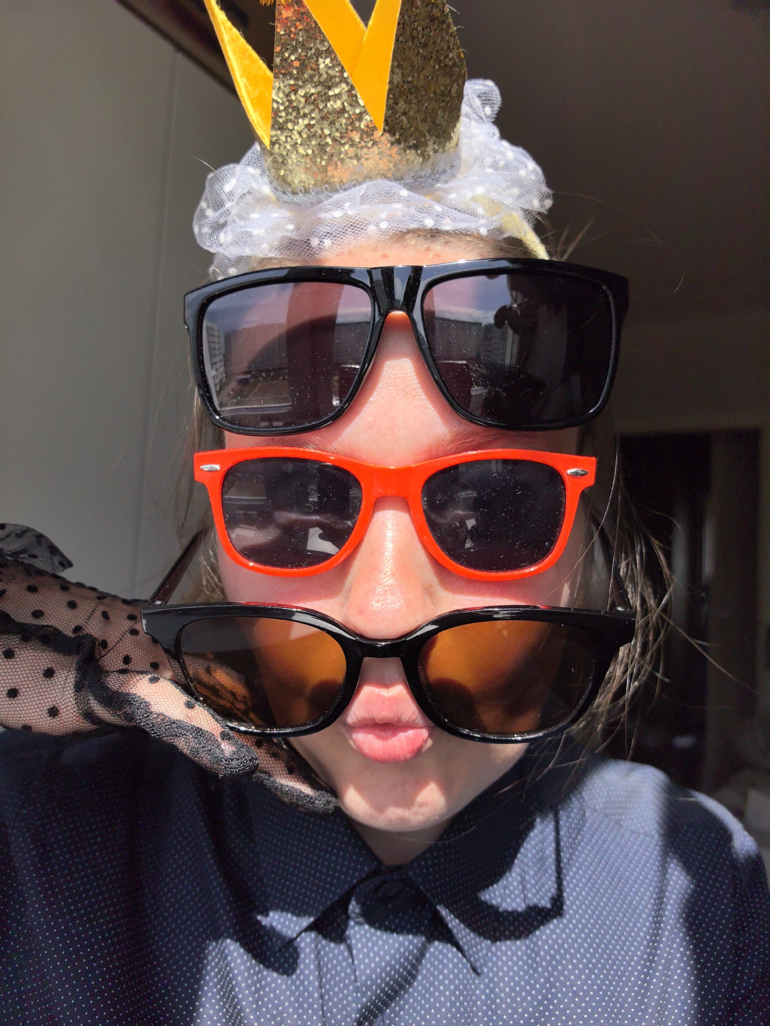 A person is holding three pairs of sunglasses stacked on their face. The top pair is black, the middle pair is red, and the bottom pair is black with amber lenses. The person is wearing a gold glitter crown and white mesh headpiece. They are also wearing a navy blue, polka-dotted button-down shirt and a black lace glove with polka dots. The location of the image is unknown.
