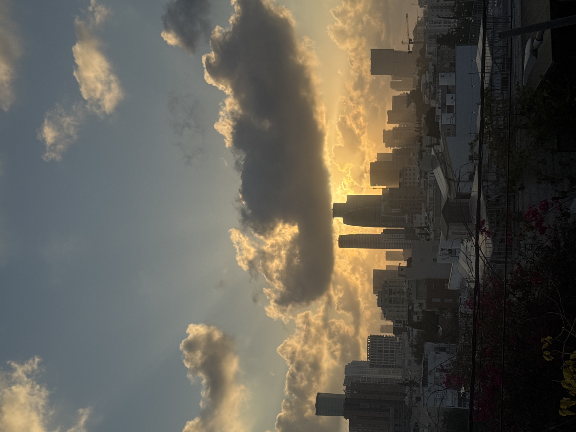 The image shows a skyline of Ramat Gan, Israel, at sunset. The sun is setting behind a dense cluster of clouds, casting a warm, golden light over the city. The sky is a gradient of blue and orange, with wispy clouds in the upper portion and darker, more dramatic clouds closer to the horizon. Several tall buildings, including skyscrapers, are visible in the distance, silhouetted against the sunset. In the foreground, there are rooftops with some greenery and hints of vegetation. The overall scene is one of urban landscape illuminated by the dramatic colors of a sunset.