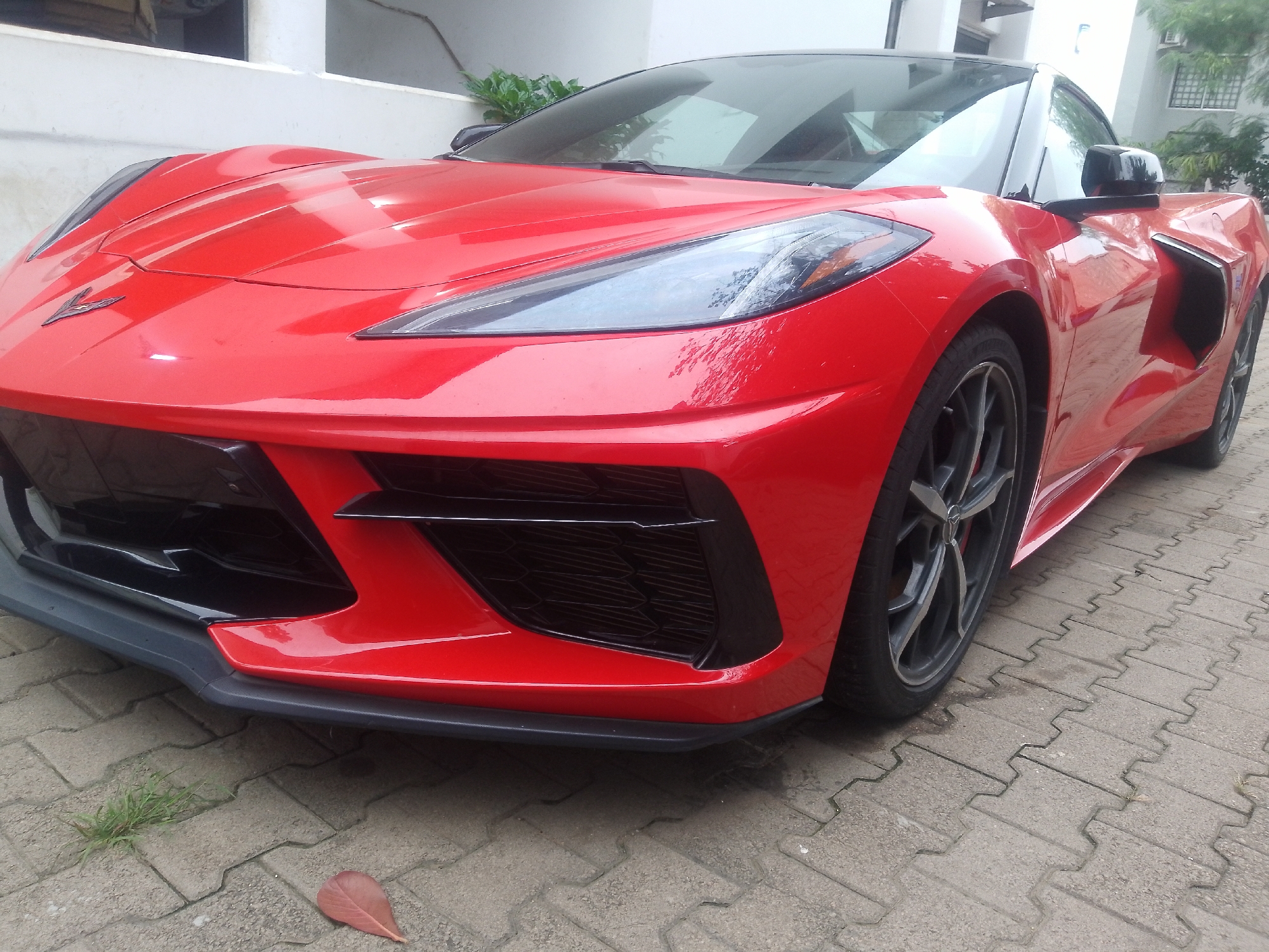 A red Chevrolet Corvette C8 is parked on a textured grey pavement in an outdoor urban setting, possibly in Abuja, Nigeria, given the style of building in the background, which features white rendered walls and dark-framed windows. The car is positioned at an angle, with the front passenger side wheel and the front bumper area prominently displayed. The car's sleek design is evident, with its sharp headlights, a large black grille, and aerodynamic lines. The pavement is made of interlocking concrete blocks, with small patches of grass and a single fallen leaf visible. In the background, the facade of a building with an upper story is visible, partially obscured by foliage. The scene appears to be a static, daytime shot with natural lighting.