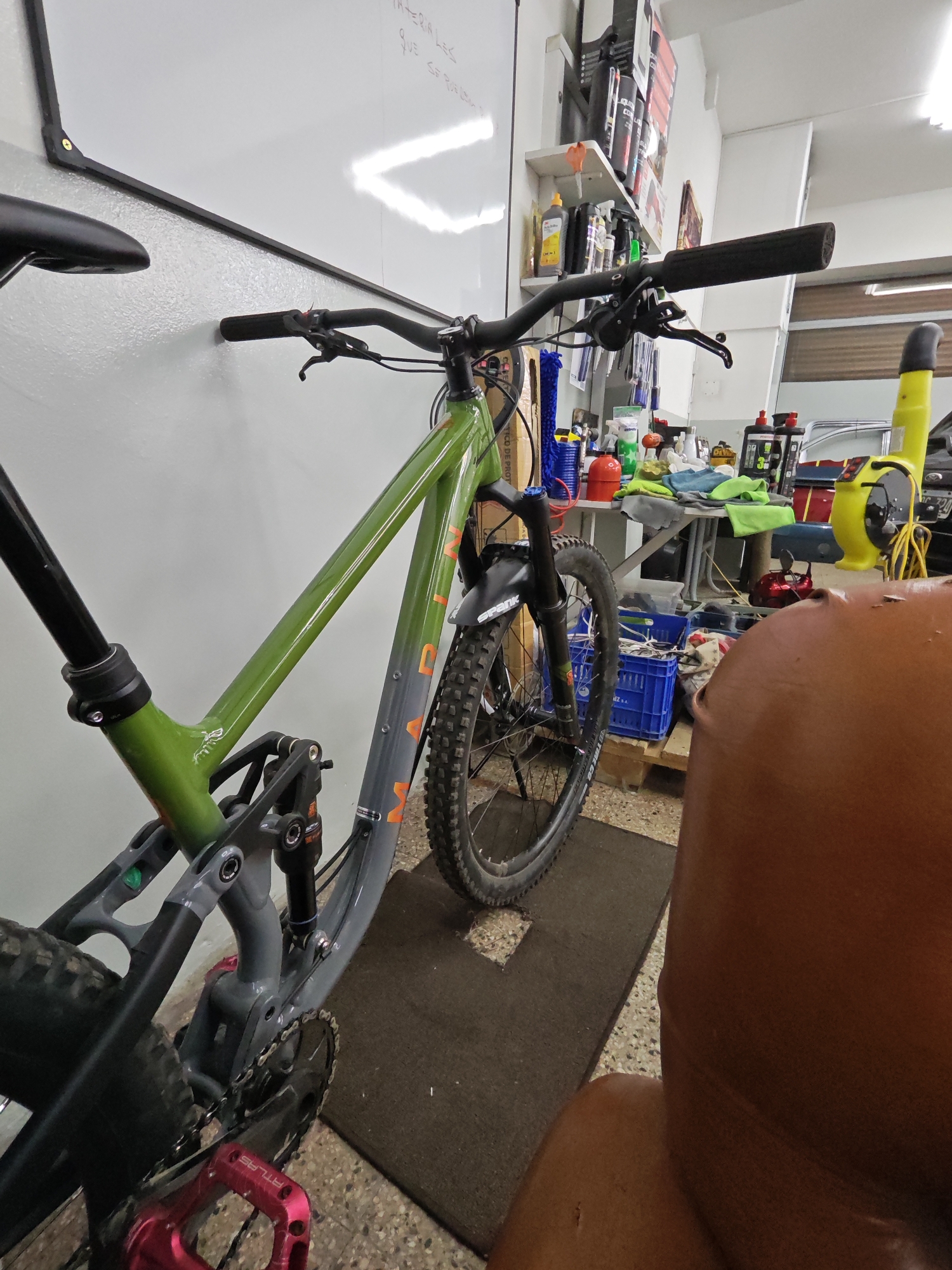 The image shows a green and gray mountain bike leaning against a white wall. The bike has a black seat and handlebars, a front suspension fork, and pink pedals. The bike is resting on a brown mat. In the background, there is a cluttered workspace with a table, shelves, tools, and various objects. The location appears to be an indoor space, possibly a workshop or garage. It's impossible to determine if the location is in Buenos Aires, Argentina without any more information. There's also part of a person visible in the front right corner of the image, out of focus.
