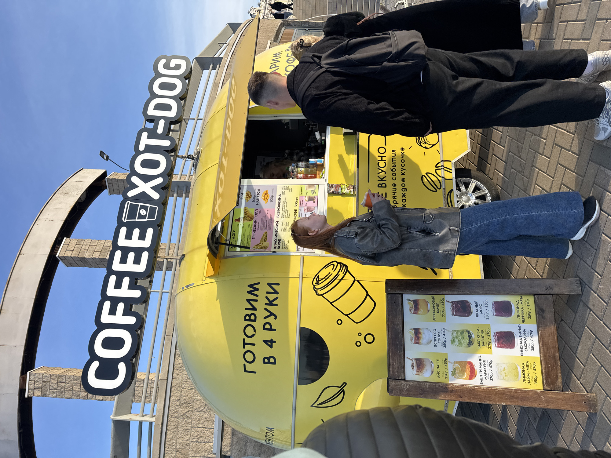The image shows a yellow coffee and hot dog food truck in Nizhny Novgorod, Russia. The truck has a large sign reading "COFFEE XOT-DOG." The sides of the truck are decorated with illustrations, including a coffee cup and a leaf. In front of the truck is a menu board listing various drinks and their prices in Russian.
A young woman with red hair stands near the menu board, holding a cup and looking up at the menu above the service window. A man in a black jacket stands beside her.