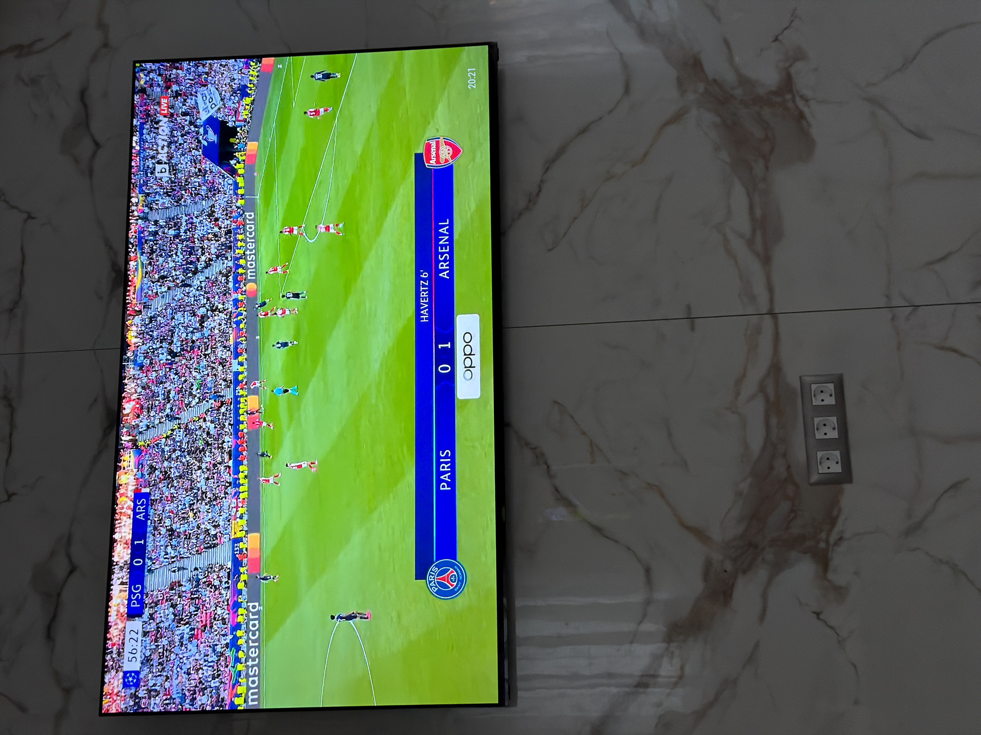 The image shows a television displaying a live broadcast of a football match between Paris Saint-Germain (PSG) and Arsenal. The score is 0-1 in favor of Arsenal, with Havertz scoring in the 6th minute. The time displayed on the screen is 56:22. The broadcast includes branding from Mastercard and Oppo. The television is mounted on a wall with a marble-like pattern. To the right of the television, there is a set of three electrical outlets. The image was likely taken indoors, potentially in a home setting. The location is not specified in the image and cannot be determined from the visual information provided.