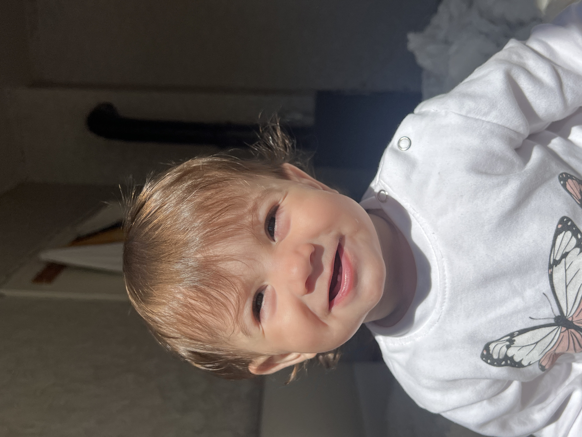 A baby with light brown hair is smiling at the camera. The baby is wearing a white long-sleeved shirt with a butterfly design on the right side. The background is a dark gray with some light patches. There is no indication of the city of Elena, Bulgaria, in the image.