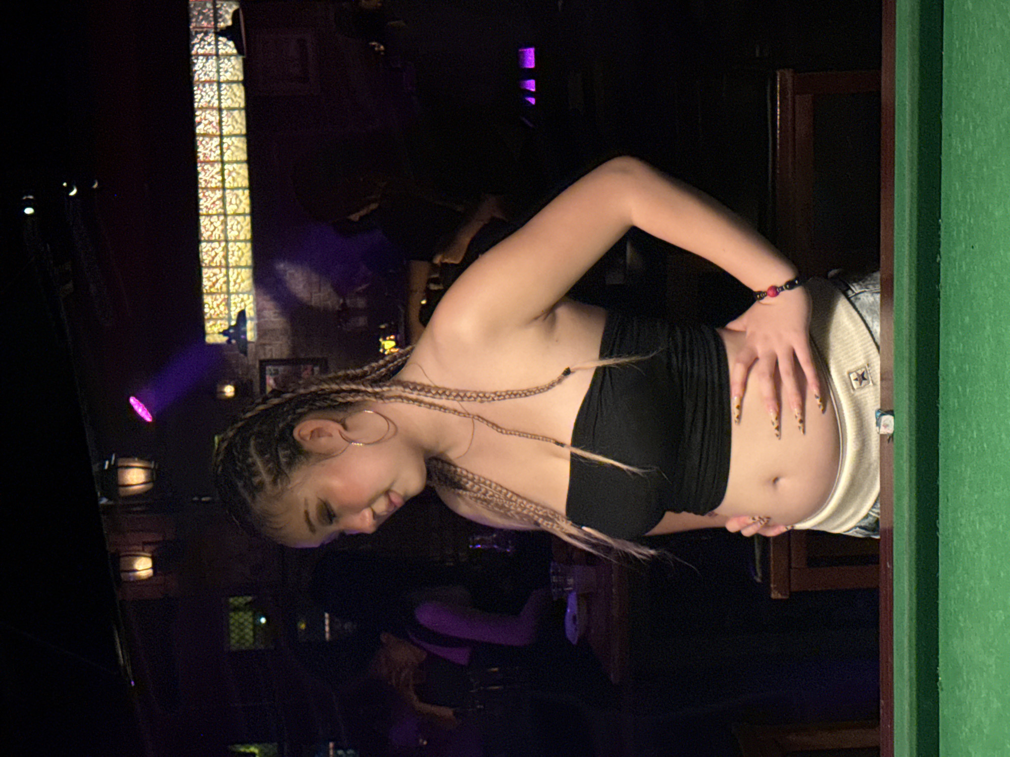 A woman with braided hair and a black tube top stands in a dimly lit bar. She is positioned in the foreground, facing slightly to the left, with her right hand resting on her hip and her left hand on her stomach. She is wearing a light-colored waistband with a star logo and denim shorts. A green pool table is visible in the lower part of the image. In the background, other patrons are dimly visible at tables, and the bar features decorative lights and lamps. The scene appears to be a casual nightlife setting. The image location is specified as Liangjiang, China.