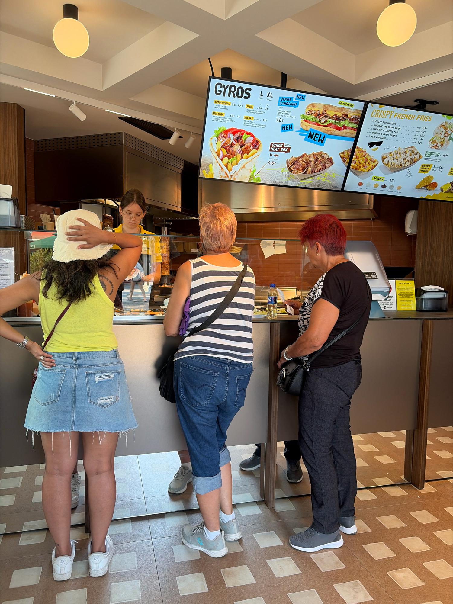 The image shows a gyros restaurant in Budva, Montenegro. Three people are visible. A woman in a yellow tank top and denim skirt stands at the counter with her back to the camera, wearing a white bucket hat. To her right, a woman with short, red hair, wearing a striped t-shirt and jeans, is looking at a menu. A third woman, with dark hair and wearing a black and white patterned shirt and dark pants, is also looking at a menu. A young woman in a yellow shirt is serving behind the counter. A digital menu board displays food items and prices. The restaurant appears clean and modern. The floor has a distinctive tiled pattern.