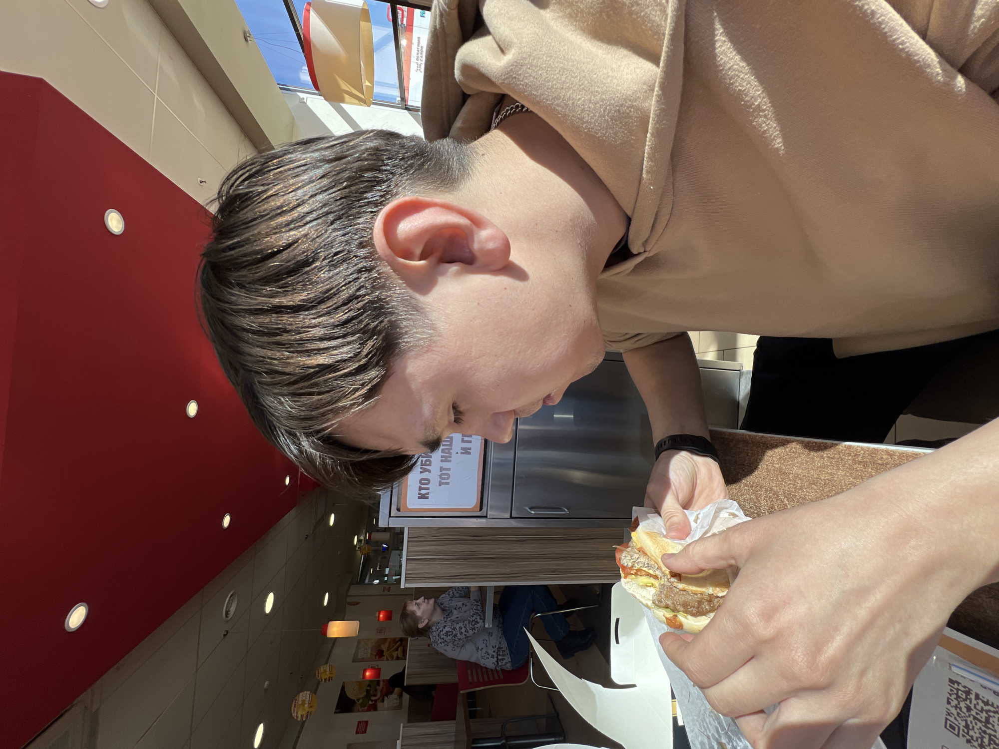 A young person, likely male, with medium-brown hair and wearing a beige hoodie, is captured in a close-up, downward-facing perspective. They are holding a burger wrapped in white paper with both hands, appearing focused on eating it. A silver chain is visible around their neck, and a black watch is on their left wrist. The burger rests in a white cardboard holder on a brown textured tabletop.
The setting appears to be a fast-food restaurant or food court in Izhevsk, Russia, indicated by the style and visible Russian text. In the background, behind the main subject's head, is a stainless steel counter. A white sign with Cyrillic text, partially visible, reads "КТО УБИВАЕТ ТОТ НАШУМЕЛ И Г." (KTO UBIVAET TOT NASHUMEL I G.). To the left, a vibrant red wall with recessed circular lights adds a pop of color to the otherwise neutral background. Above, a light-colored ceiling features more recessed lighting and a skylight, revealing a clear blue sky and allowing bright natural light into the space. Decorative, rectangular light fixtures hang from the ceiling. In the distant background, another person, possibly female, is seated at a table. A QR code is visible on the table surface to the right. The bright natural light suggests it is daytime.
