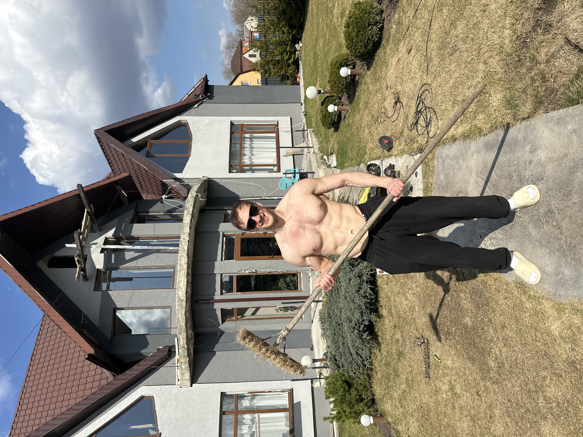 A shirtless man stands on a concrete pathway holding a rake, wearing black sweatpants, sunglasses, and white slippers. Behind him is a gray two-story house with brown trim and a brown roof, under a cloudy sky. The yard has patches of green and dry grass, bushes, and lawn lights. The man is smiling. The exact location in the city of Kursk, Russia, cannot be determined from the image.