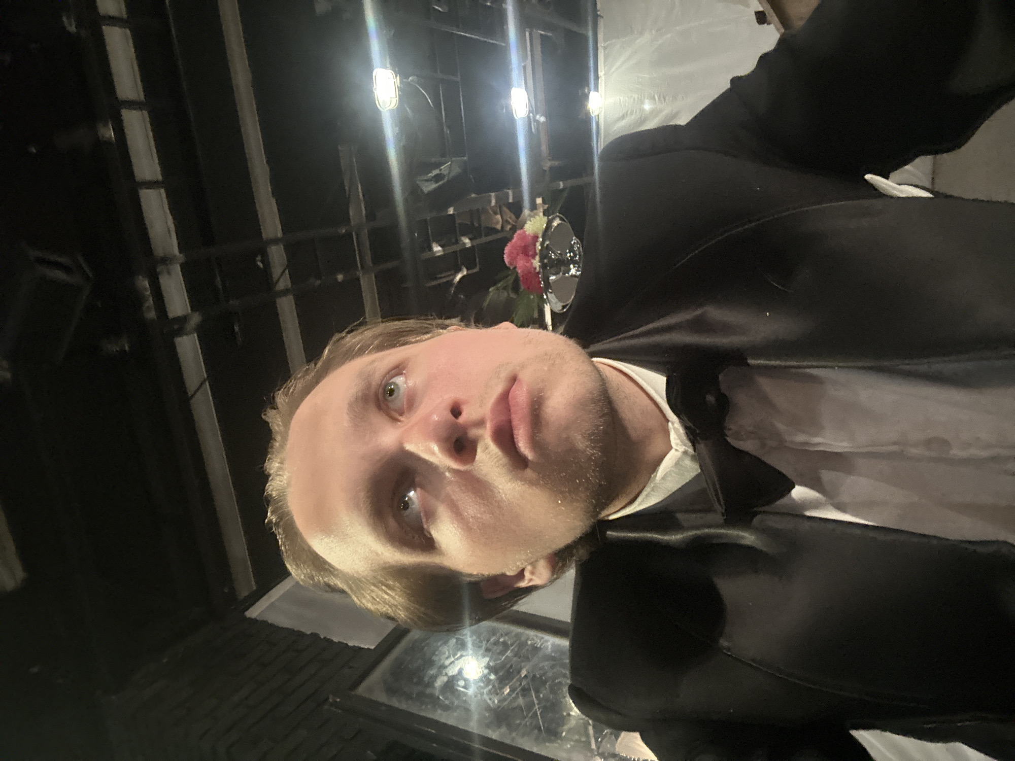 The image is a low-angle, close-up, selfie-style shot of a young man wearing a tuxedo with a bow tie and white shirt. He has light brown hair and is looking upwards. The background appears to be an indoor venue with exposed ceiling structures, lights, and some white sheeting or fabric. A small floral arrangement with pink and white flowers is visible to the right of the man. The overall lighting is dim with some bright highlights from the overhead lights. The location is specified as Unknown, Unknown.