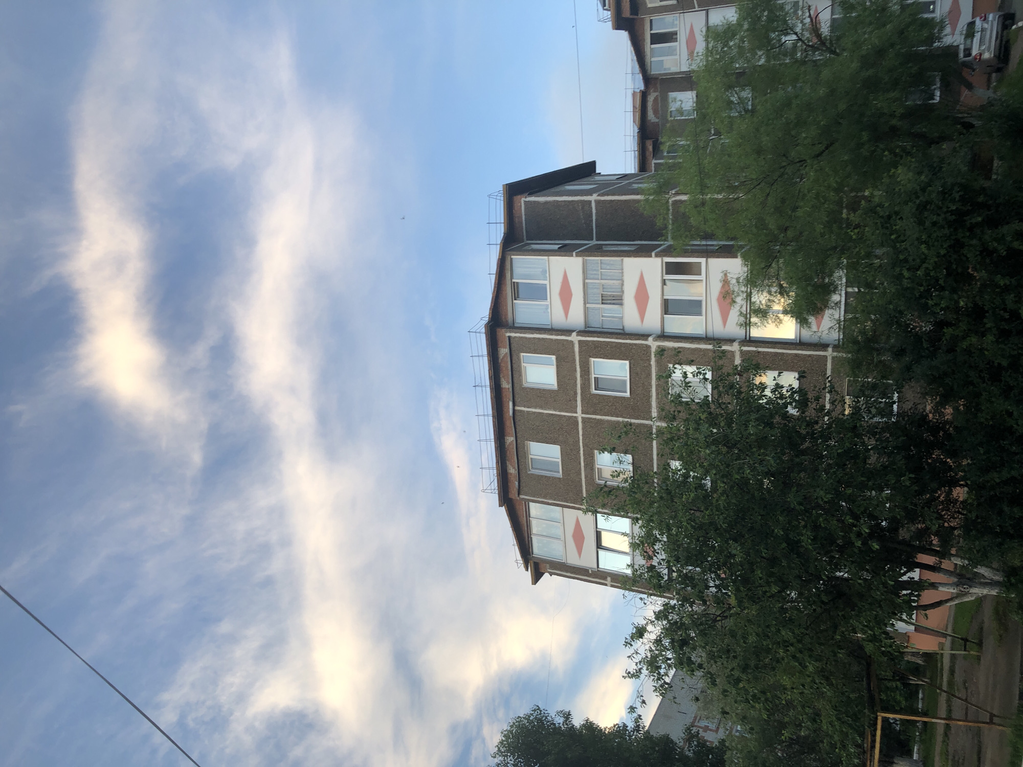 The image shows a view of a concrete apartment building in Solikamsk, Russia. The building has multiple floors and windows, with a prominent diagonal design element on the facade. Trees and foliage are visible in the foreground, partially obscuring the lower levels of the building. The sky above is blue with wispy white clouds, suggesting it is daytime. A power line stretches across the upper portion of the frame. The scene depicts a residential area in the city.
