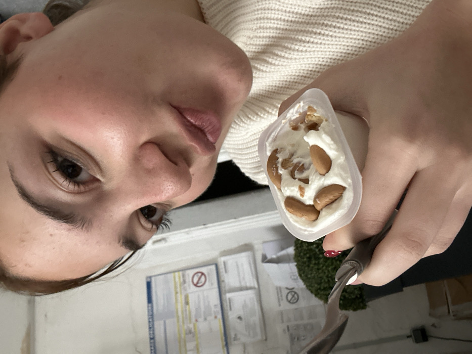 The image presents a close-up, indoor scene. A female subject, visible from the upper chest upward and oriented towards the upper right, has fair skin, dark eyebrows, visible eyelashes, and her lips are slightly puckered. She is wearing a cream-colored ribbed knit sweater. Her left hand, with red and white polka-dotted fingernails, holds a clear plastic cup containing a white creamy substance, topped with several small, oval, light-brown cookies and cookie fragments. A silver-colored metal spoon with a dark handle is also held in her left hand, positioned near the cup. The background to the left of the subject features a white wall with multiple papers affixed to it. One visible notice contains blue text and a red circular 'no' symbol over an icon, with the partially legible French word "OBLIGATOIRE" indicating mandatory information. Below these notices, a dark planter holds a spherical green plant. This indoor setting, marked by the French text "OBLIGATOIRE," is located within a building in Strasbourg, France, possibly a breakroom or communal area.