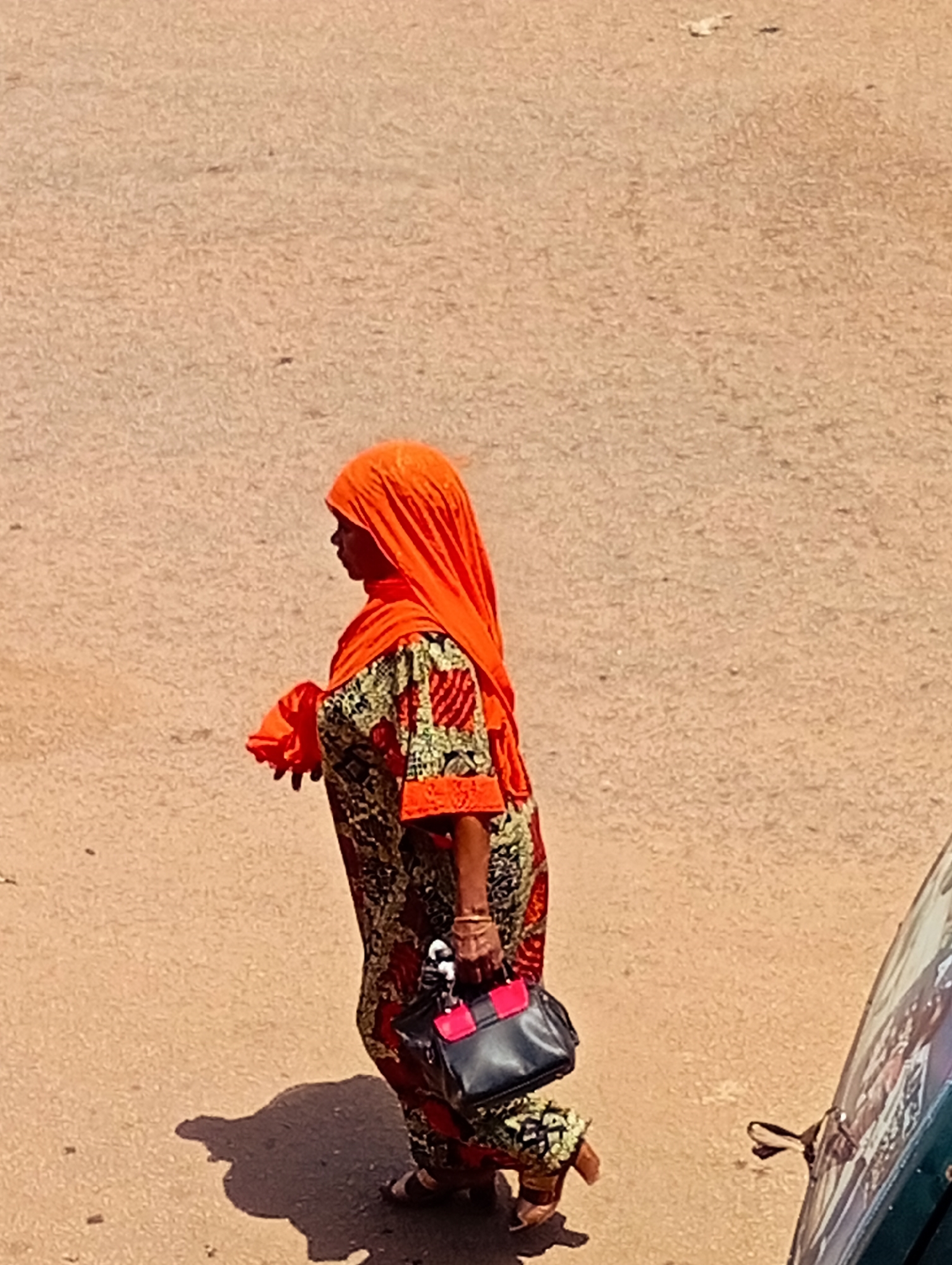 A person wearing a bright orange hijab and a patterned dress walks on a sandy road in Bukuru, Nigeria. The person is carrying a dark bag with pink accents. The ground is a light brown color, and there is a shadow cast on the road. Part of a green vehicle is visible on the right side of the image.