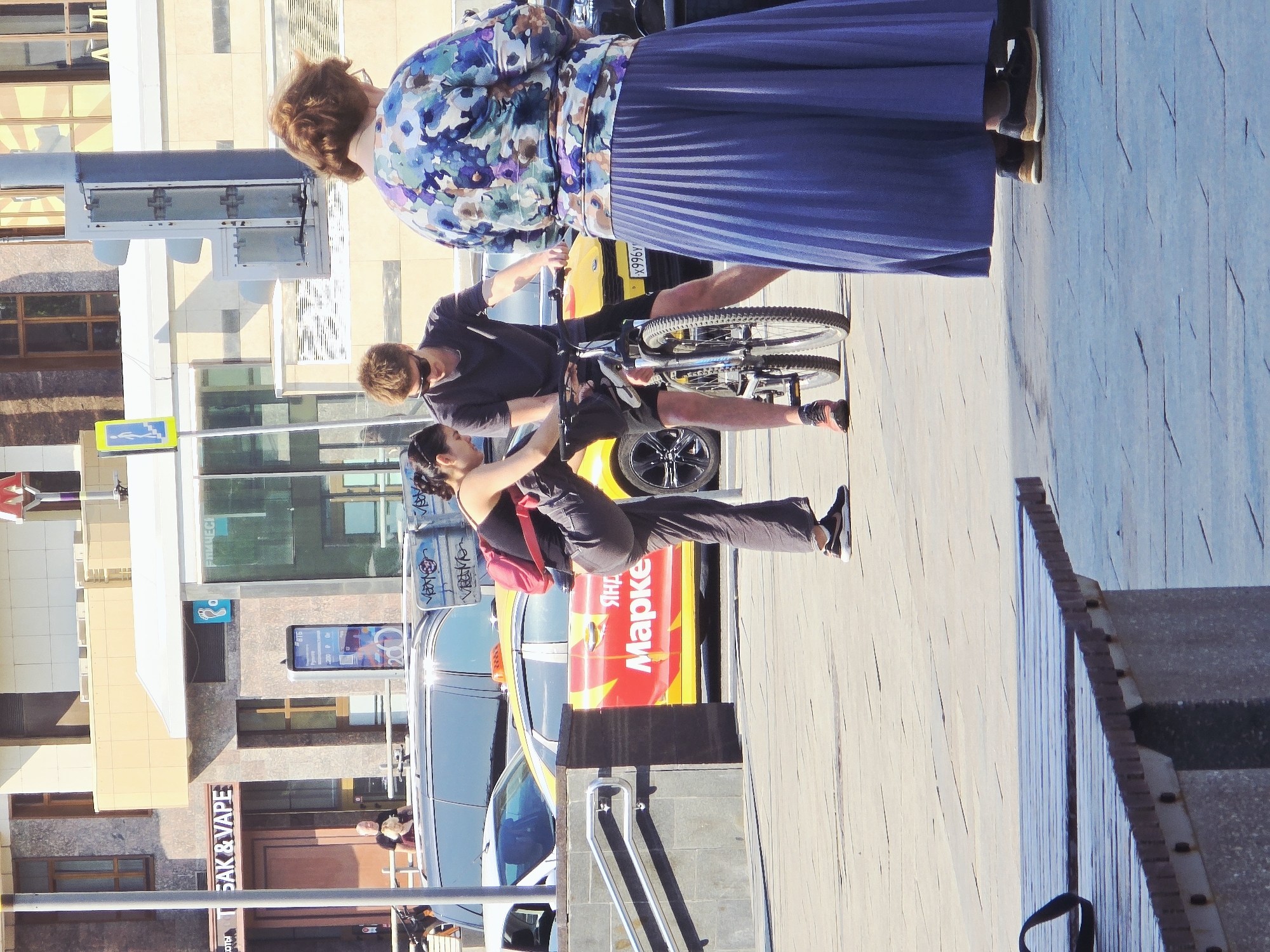 Here is a description of the image:
The image captures a street scene in Moscow, Russia. Three people are prominently featured: an older woman with auburn hair wearing a floral print top and long pleated blue skirt, a younger man with light brown hair wearing a dark shirt and shorts who is on a bicycle, and a young woman with dark hair in a tank top, dark pants, and a small red backpack. The young woman is seemingly handing something to the man on the bicycle.
The setting includes elements of city infrastructure such as buildings with glass facades, signage, and street-level shops. A yellow taxi cab with a red side panel displaying Cyrillic script is visible behind the group. There's also a pedestrian crossing sign and a glimpse of an establishment with Cyrillic lettering. The scene takes place on a sidewalk with a textured surface.