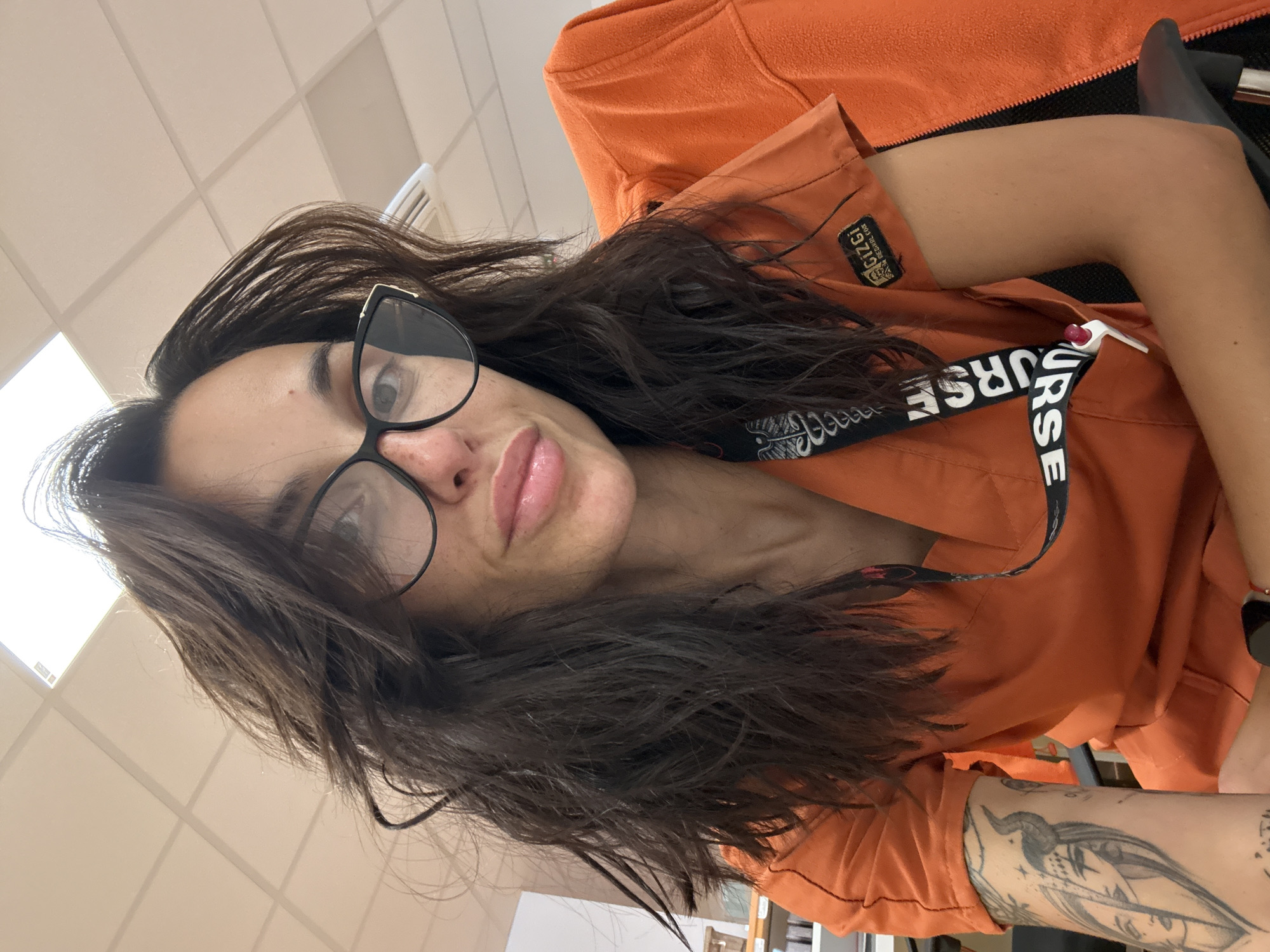 A female subject is depicted in a close-up selfie taken indoors in Pleven, Bulgaria. She has long, dark, wavy hair and wears black-framed eyeglasses with gold accents on the outer corners. Freckles are visible on her nose and cheeks, and her lips appear plump and glossy. She is wearing an orange V-neck medical scrub top with a dark brown "GIZGI MEDICAL OR" patch on the left sleeve. A black and white lanyard, repeatedly printed with "NURSE", is around her neck, from which a white penlight is clipped. A prominent black and grey tattoo, depicting a horned female face and other intricate designs, covers her left forearm. An orange fleece jacket is draped over an unseen chair to her right. The background features a dropped ceiling with square acoustic panels, integrated fluorescent lighting, and a white air vent in the upper right quadrant.