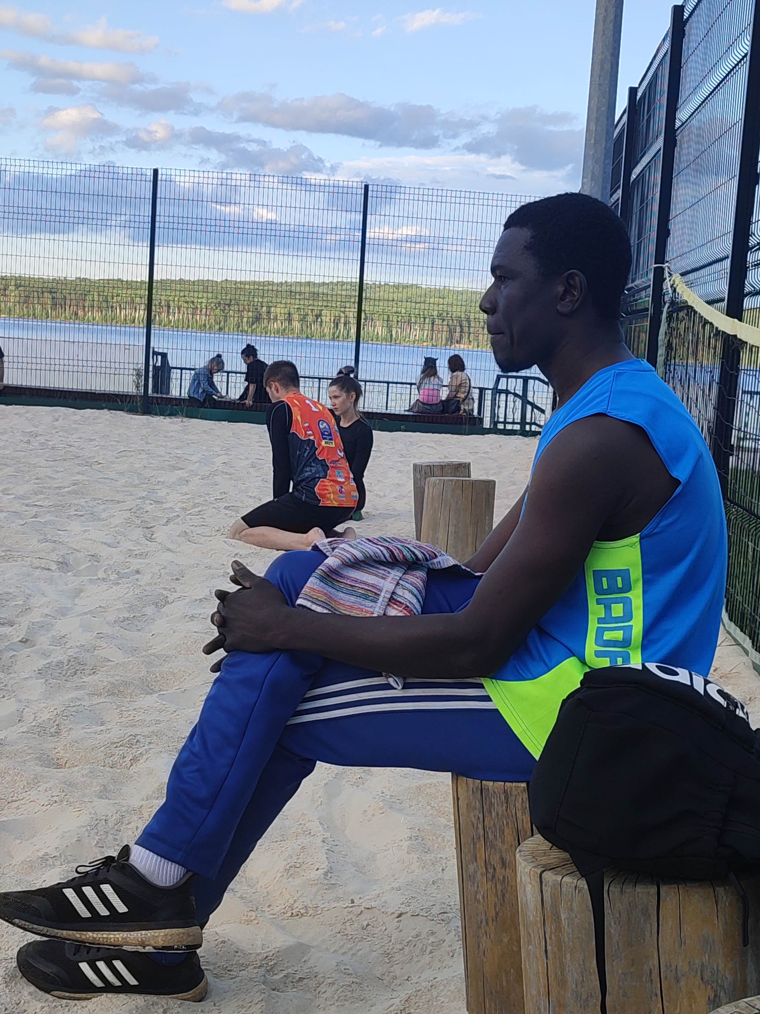In a sandy outdoor setting, likely a beach volleyball court, a Black man is seated on two wooden logs. He wears a blue and neon green sleeveless athletic shirt with "BAD" visible in neon green lettering down the side. His blue athletic pants have white stripes. On his feet are black athletic shoes with white stripes. Next to him on the logs is a black backpack.
In the background, through a fence, a body of water and a tree line are visible. Other people are scattered on the sand behind the main subject. One person in the mid-ground wears an orange and black athletic jersey. The sky is partly cloudy, suggesting it is daytime, possibly late afternoon or early evening. The overall mood appears relaxed.