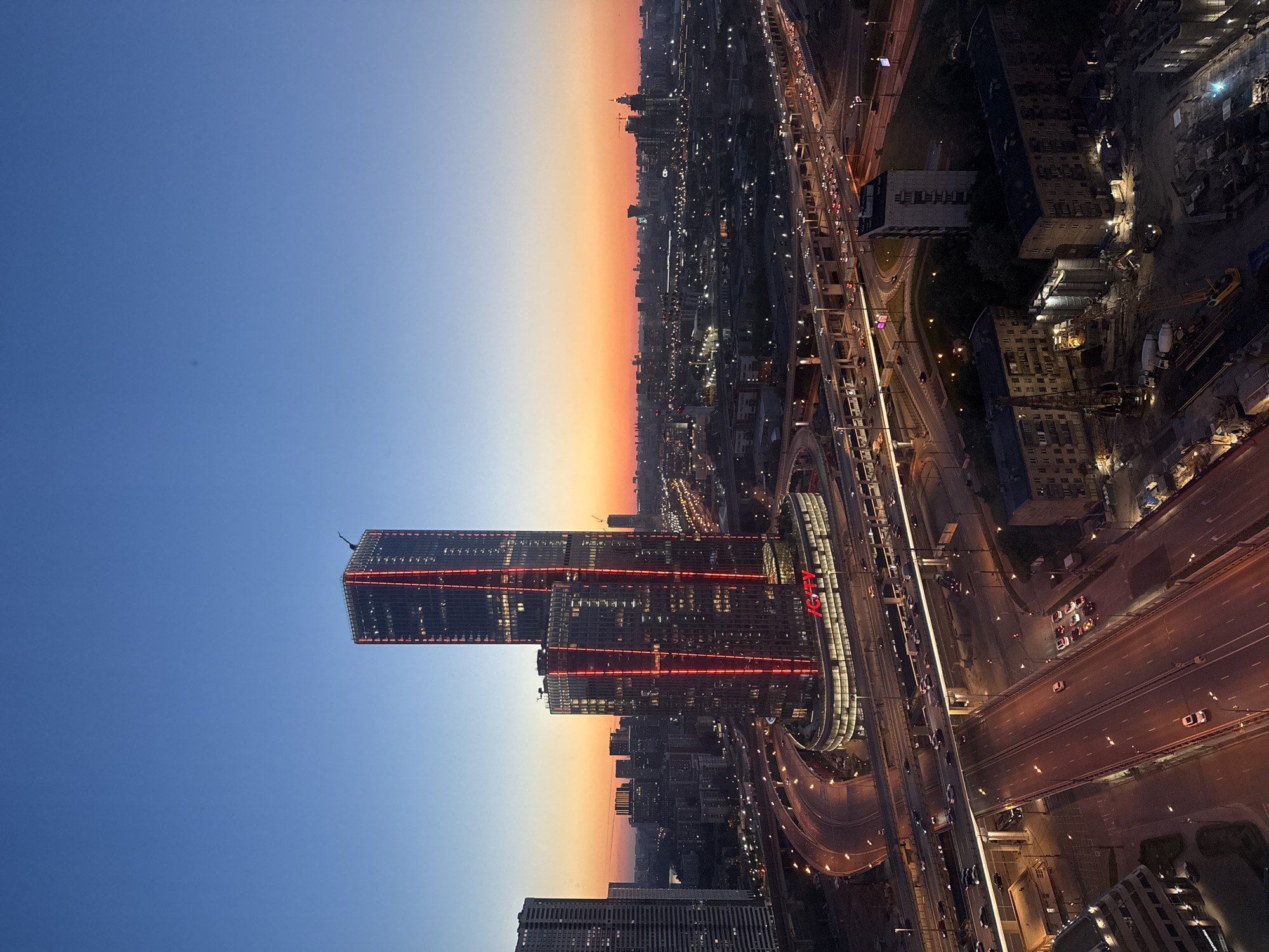 This high-angle, wide shot depicts a sprawling urban landscape at dusk, with the horizon glowing in shades of orange and pink transitioning to deep blue overhead. The scene is dominated by two modern skyscrapers, part of the "I CITY" complex, prominently featuring vertical red LED lighting strips along their facades and an illuminated "I CITY" sign at their shared base. These buildings are located within the Moscow International Business Center (Moscow-City), Moscow, Russia.
Below the skyscrapers, a complex, multi-level highway interchange system with numerous lanes, on-ramps, and off-ramps stretches across the midground. Vehicle headlights and taillights form glowing lines on the asphalt, indicating active traffic flow. Streetlights illuminate the roads and surrounding areas.
To the left of the skyscrapers, another tall, less illuminated residential building is visible. In the foreground, below the highway, there is an active construction site with several large cranes, excavators, cement trucks, and concrete silos, illuminated by bright work lights. Adjacent to the construction site are several older, multi-story residential buildings, some appearing partially obscured by trees. The cityscape extends into the distance, revealing a dense network of buildings and illuminated streets under the twilight sky. No specific individuals are discernible, but the presence of numerous vehicles implies human activity. A faint, small, dark anomaly is visible in the upper left quadrant of the sky.