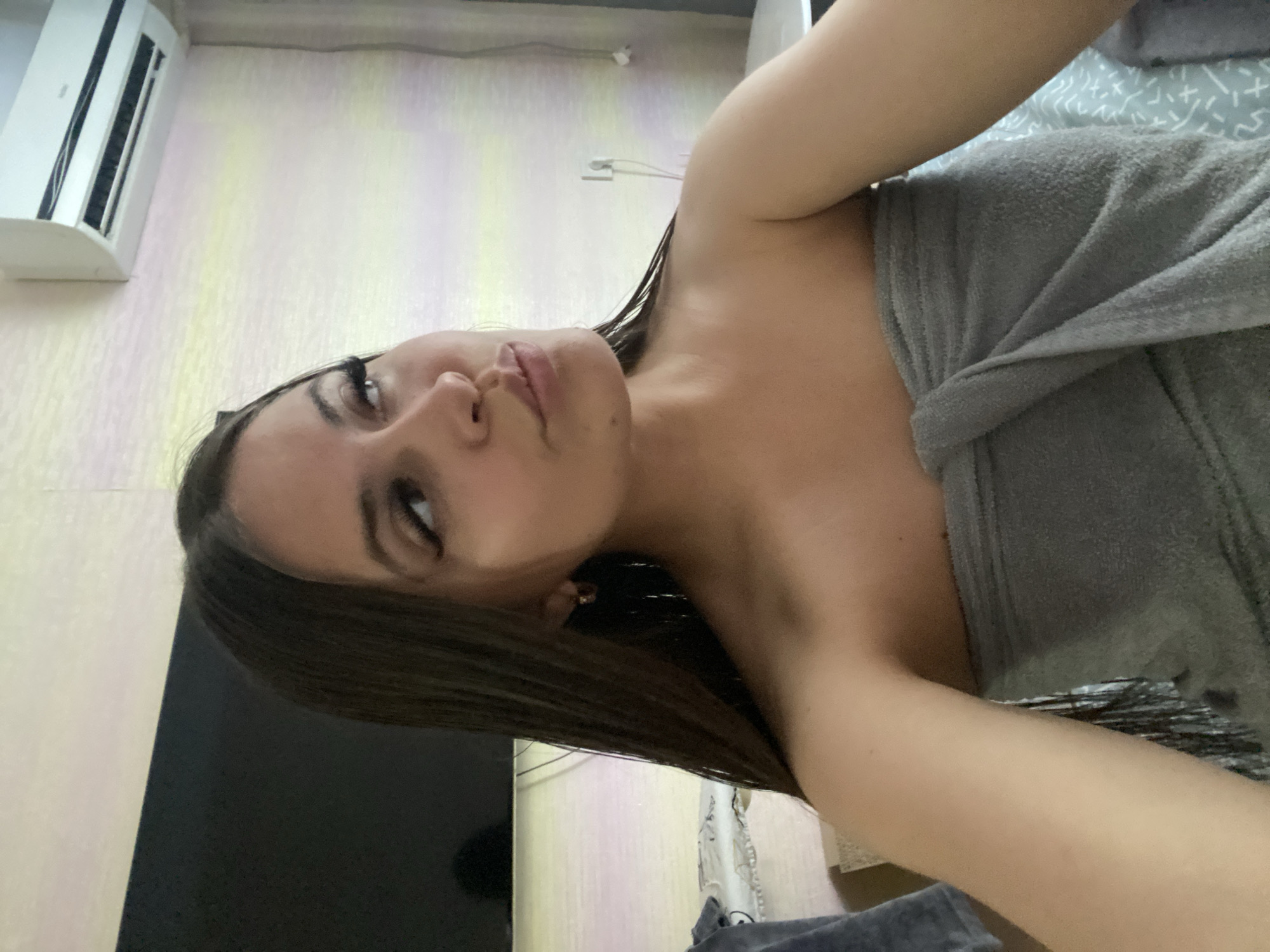 The media file is a selfie taken by a young woman in Makhachkala, Russia. She is wrapped in a grey towel, suggesting she may have just showered or is getting ready. Her dark hair is wet and falls around her shoulders. She is looking upwards towards the camera with a slightly playful expression, her eyes are made up with mascara. The background appears to be a room with light-colored walls, one of which has a subtle striped pattern. An air conditioning unit is visible on the wall in the upper left corner of the frame. A dark object, possibly a television, is visible in the lower left. The lighting suggests it is daytime, though no external weather cues are present.