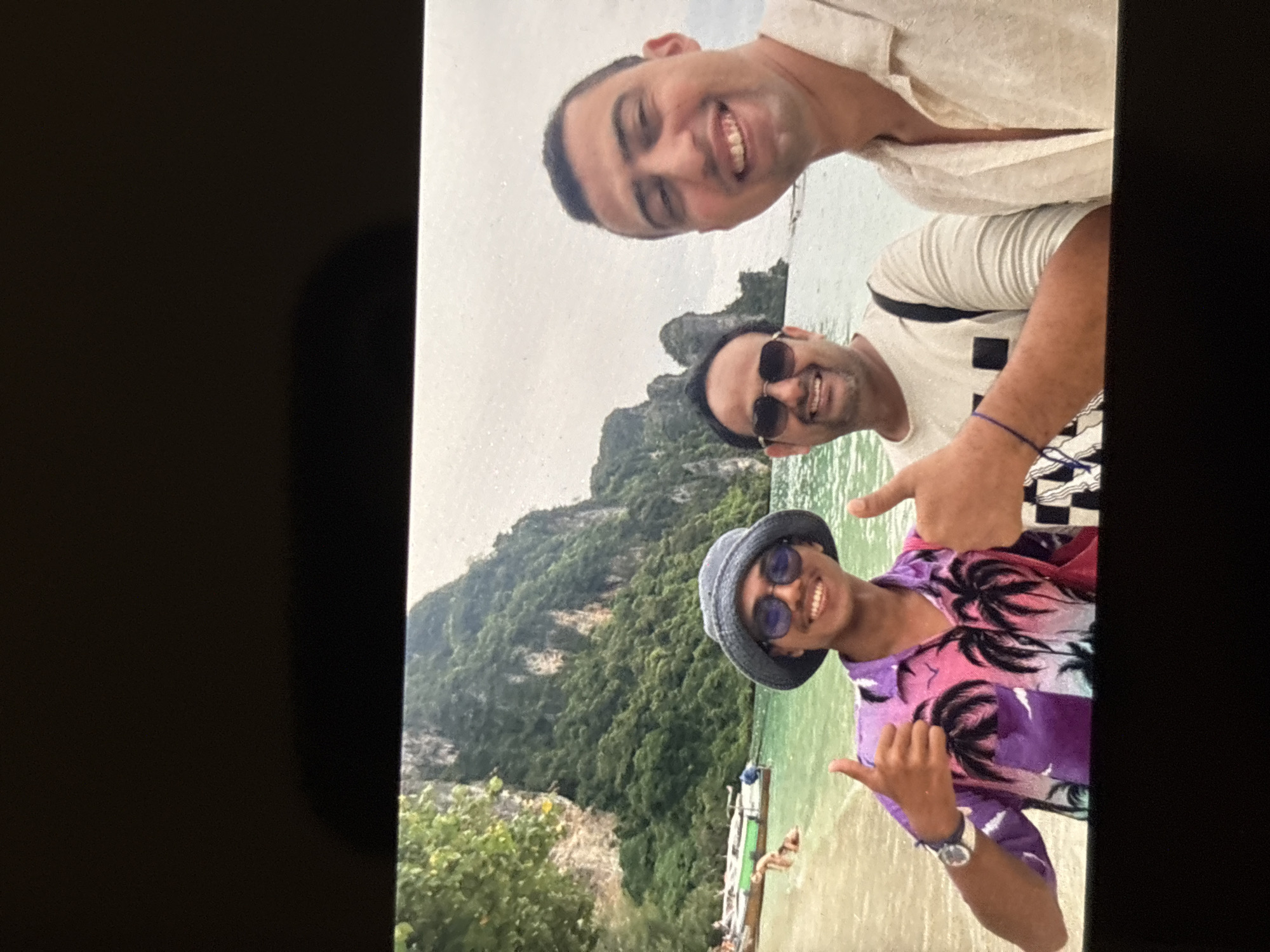 Three men are taking a selfie on a beach with large limestone cliffs in the background. The man on the left is wearing a purple and pink short-sleeved shirt with a palm tree pattern, a blue bucket hat, and sunglasses. He is giving a thumbs-up with both hands. The man in the middle is wearing a light-colored long-sleeved shirt and sunglasses, and he is smiling. The man on the right is wearing a light-colored short-sleeved shirt and is also smiling. The water is a clear turquoise color, and there are longtail boats pulled up on the sandy beach. This scene is consistent with popular tourist locations in Chalong, Thailand, such as Ao Maya or a similar bay. The overall mood is one of leisure and enjoyment.