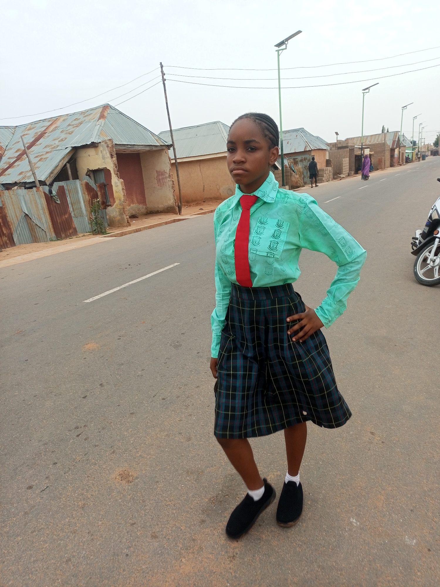A young girl stands on a paved road in Tempure, Nigeria. She wears a light green long-sleeved shirt, a red tie, a plaid skirt, and black shoes with white socks. She has dark hair braided back from her face. Her left hand is on her hip, and she looks directly at the camera with a neutral expression.
Behind her are several simple, single-story buildings with corrugated metal roofs. Some buildings are painted with fading earth tones and show signs of disrepair. Utility poles line the road. In the background, other people can be seen walking along the street. A motorcycle is parked on the right edge of the frame. The sky is overcast. A white lane marker is visible on the road.