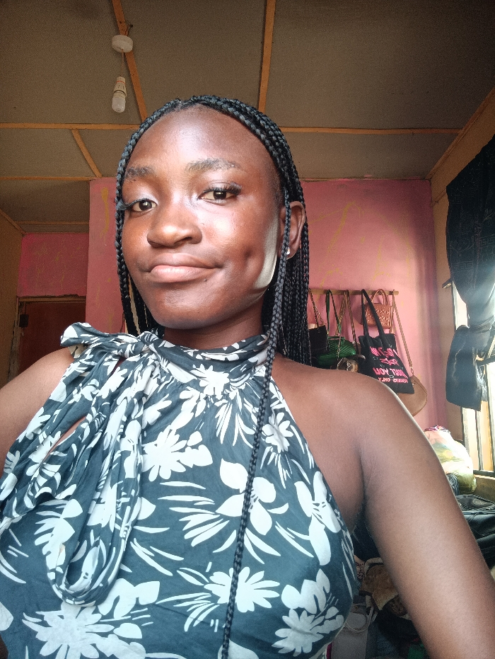 A young adult female is depicted indoors, located in the city of Oye, Nigeria. She has dark skin, black hair styled in numerous small braids, and prominent dimples visible on both cheeks. She looks directly at the camera with a subtle smile. She wears a halter-neck top with a dark blue-green base adorned with white floral patterns; a knotted tie is visible on the left shoulder area. A silver-colored hoop earring is present in her right ear. The indoor environment features a ceiling constructed from light brown wooden panels and beams, from which a single, opaque white light bulb hangs by a white cord. The wall behind the subject is painted a faded pink. To the right, several bags of various designs are suspended against the wall, with an obscured opening or window beyond. A section of a dark brown door or doorframe is visible on the left side of the frame.