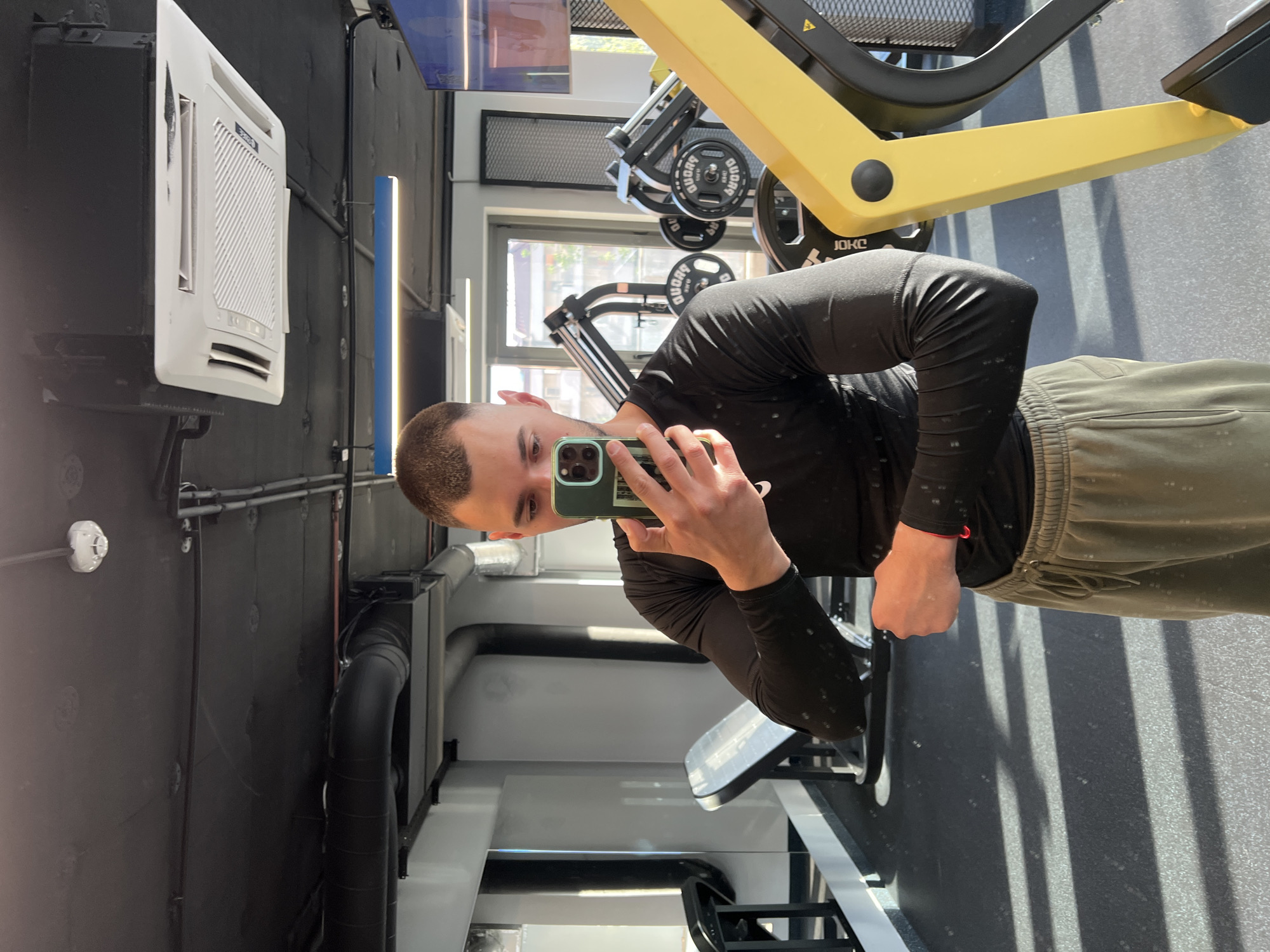 A young man is captured in a selfie taken inside a gym, holding a smartphone with a green case. He appears to be looking into the camera with a neutral to slight smile. He has short, dark hair and is wearing a black long-sleeved athletic shirt and olive green sweatpants. His left arm is bent, and his fist is clenched.
The gym interior features dark, possibly black, walls and ceiling, contrasting with the bright natural light entering from a large window. This sunlight casts strong, defined shadows on the grey, speckled floor, suggesting it is daytime. Exposed black pipes and ventilation ducts are visible along the walls and ceiling, contributing to an industrial aesthetic.
Various gym equipment can be seen. A prominent yellow leg press machine is in the mid-ground. Black weight plates, some marked with "DUGAR GYM" and "JOKO", are stacked on equipment. In the background, other gym machines are partially visible. A white air conditioning unit, possibly from the brand "Gree" (partially visible), is mounted on a dark wall. A blue vertical light strip is also visible in the background near a window. The overall environment suggests a modern fitness facility in Plovdiv, Bulgaria, as per the provided context.