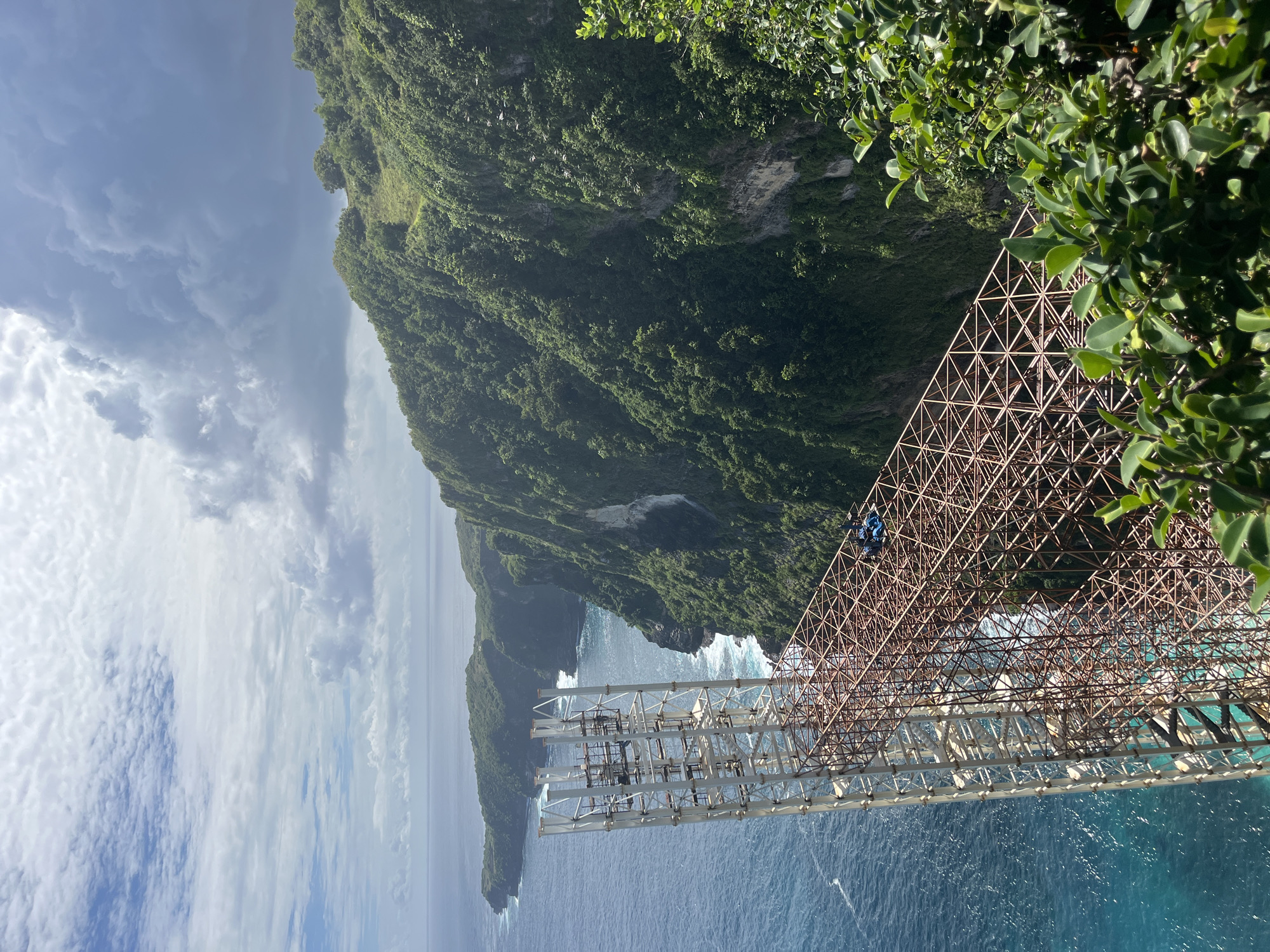 This vertical image, when rotated 90 degrees clockwise, presents a breathtaking panoramic view of the dramatic coastline of Nusa Penida, Indonesia, captured from a high vantage point. The scene is dominated by a massive, intricately designed metal structure extending from a lush green cliff face over the vibrant turquoise ocean.
In the foreground and midground, a complex lattice-work of brown, rust-colored metal beams forms what appears to be a large bridge or viewing platform. Further along this structure, it transitions into lighter, possibly white, metal sections that support smaller, scaffold-like components. One person, clad in blue clothing, is visible on the brown metal framework, suggesting ongoing construction, maintenance, or inspection activity.
To the right, the cliff face rises steeply, densely covered in rich green vegetation and trees, showcasing the island's tropical environment. Patches of lighter rock are visible through the foliage. In the distance, beyond the metal structure and across the expanse of the deep blue ocean, more rugged green cliffs and a small islet are visible, with white waves crashing against their bases.
The sky above is a mix of bright and overcast conditions, with fluffy white clouds scattered among darker, heavier cloud formations, indicating dynamic weather. The bright daylight suggests it's daytime, likely midday or early afternoon. The overall impression is one of grand natural beauty juxtaposed with significant human infrastructure, all set against the stunning backdrop of Nusa Penida's iconic landscape. No visible text is present in the image.