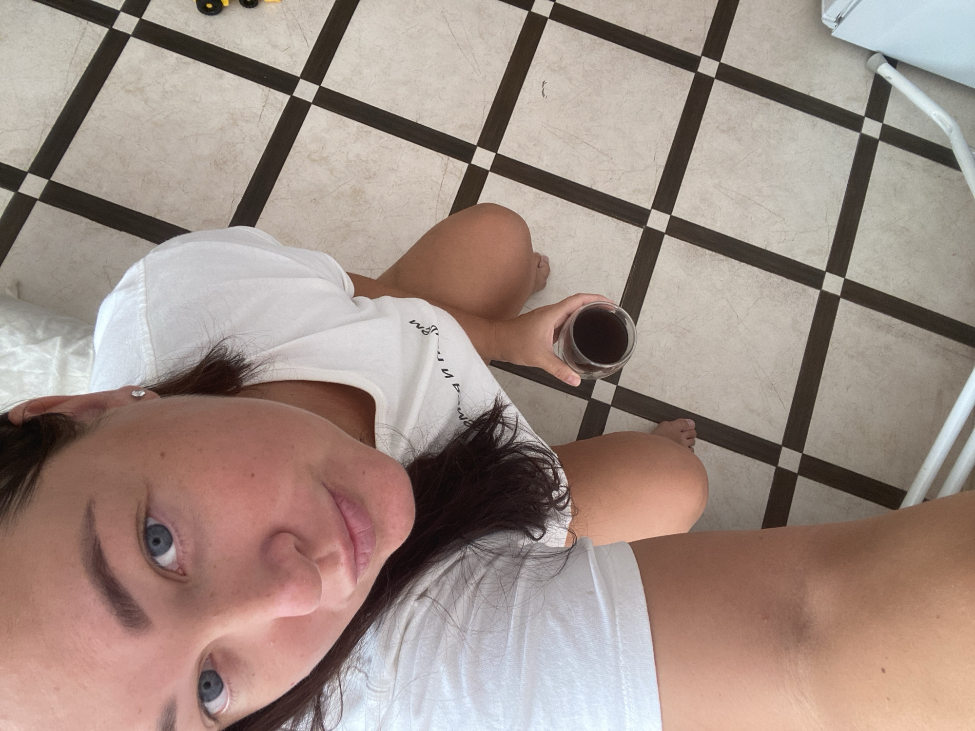 A woman with dark hair and blue eyes kneels on a tiled floor, holding a glass of dark liquid. She is wearing a white t-shirt. The floor is patterned with cream-colored tiles and dark brown grout lines forming a checkerboard. To the right of the woman, a portion of a white washing machine is visible. The scene appears to be indoors, likely in a residential setting. There is no indication of the specific location within the city of Sochi, Russia, nor any interaction or action beyond the woman's pose and grip on the glass.