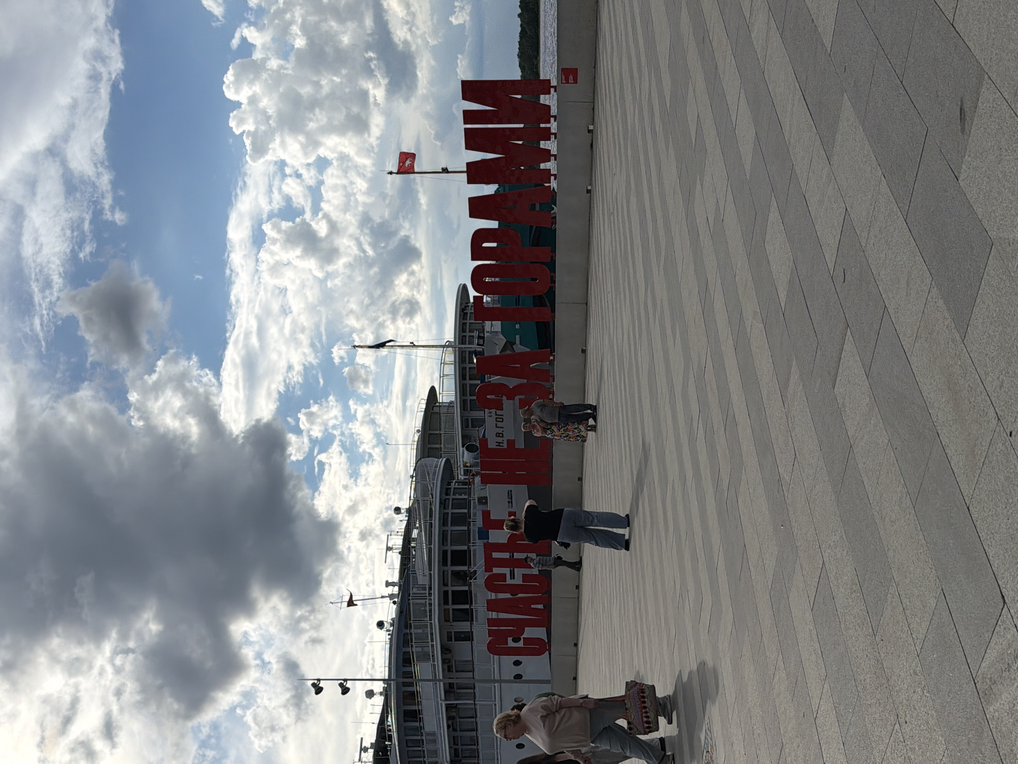 A wide shot of a waterfront promenade in Perm, Russia, features a large, red, three-dimensional Cyrillic inscription that reads "СЧАСТЬЕ НЕ ЗА ГОРАМИ" (Happiness is not far away). The inscription is positioned in front of a moored river cruise ship. Several individuals are visible on the promenade; in the foreground to the left, a woman with blonde hair and glasses walks past carrying a patterned tote bag. In the middle ground, a woman in a black top and blue jeans faces away from the camera, appearing to take a photo. Further to the right, a couple stands near the inscription, the woman in a patterned dress and the man in a grey shirt and hat. The promenade is paved with large, rectangular grey stone tiles laid in a repeating pattern. The sky above is partly cloudy with a mix of dark and bright clouds. The river is visible in the background to the right of the inscription.