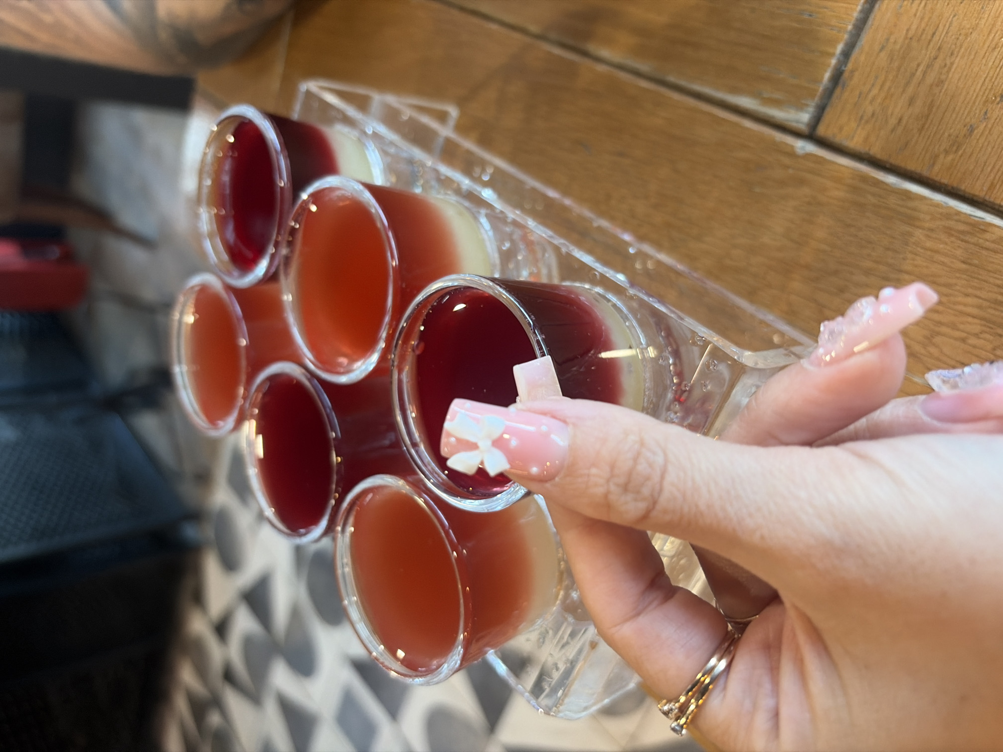 The image depicts an indoor establishment, likely a bar or restaurant, situated in Saint Petersburg, Russia. A person's right hand, adorned with two thin rings on the ring finger, holds a clear shot glass containing a two-layered liquid. The fingernails are manicured, featuring a pink gradient with white tips, small white polka dots, and a white bow appliqué on the index finger. Six other identical shot glasses, mostly filled with the same dark red/magenta top layer and opaque off-white/cream bottom layer, are arranged on a clear, rectangular acrylic tray in the foreground and mid-ground. The tray rests on a light brown wooden table composed of visible planks. The background is blurred, revealing a distinctive floor with a geometric pattern of dark gray shapes against a lighter background. Further back, out-of-focus elements include dark furniture, a dark wall, and a tattooed arm on the far right, suggesting the presence of other individuals.