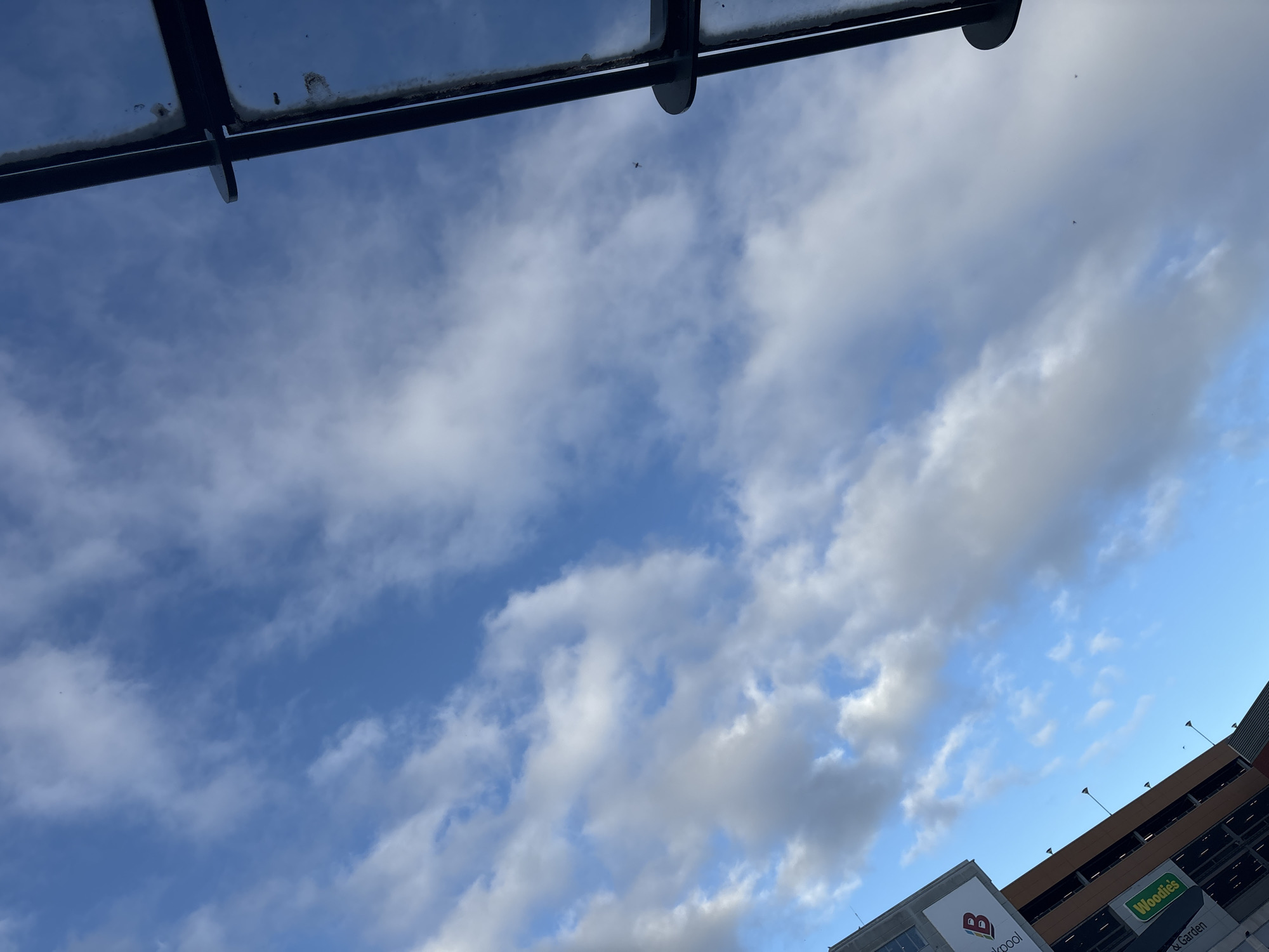 The image captures a view of a bright blue sky with scattered white clouds, taken from a low angle. In the upper right portion of the frame, a dark, angular structure, possibly part of a building or canopy, intrudes into the scene. Along the left side of the image, a multi-level building is visible, identified by signage including a large "B" logo and the text "Woodies". This building appears to be a car park, with multiple levels of open-air parking visible. The scene suggests an outdoor setting in a built-up area, with the sky dominating the upper portion of the composition. The building with the "Woodies" signage is located in Cork, Ireland.