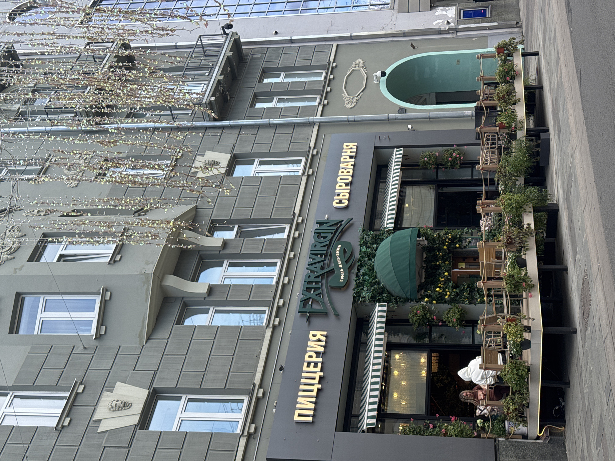 A ground-level, medium shot depicts the exterior of a restaurant named "Syrovarnya" in Moscow, Russia. The building's facade is painted a neutral grey, with architectural details featuring blocky, geometric patterns, windows, and decorative elements. The restaurant signage is vertical and uses the Russian language.
The establishment has an outdoor seating area with chairs and tables. Lush plants and flowers adorn the restaurant’s exterior, adding a touch of greenery. The storefront includes a green awning and striped green and white sunshades.
Two individuals are seated at one of the outdoor tables. The environment appears to be an urban street.