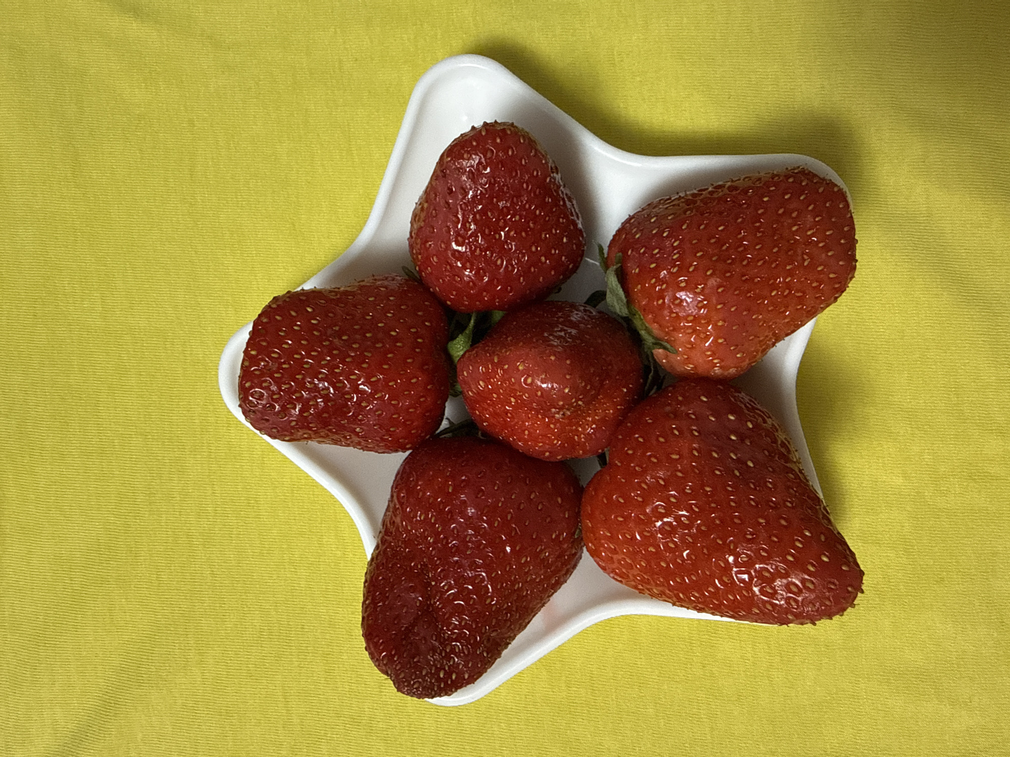 A top-down view depicts six strawberries arranged in a white, star-shaped dish. The strawberries are various sizes and shades of red, with small seeds visible on their surfaces. The dish is positioned on a yellow textured surface, possibly a tablecloth or fabric. The image does not contain any people or actions. There is no indication of this photo being taken in Chaikovsky, Russia.
