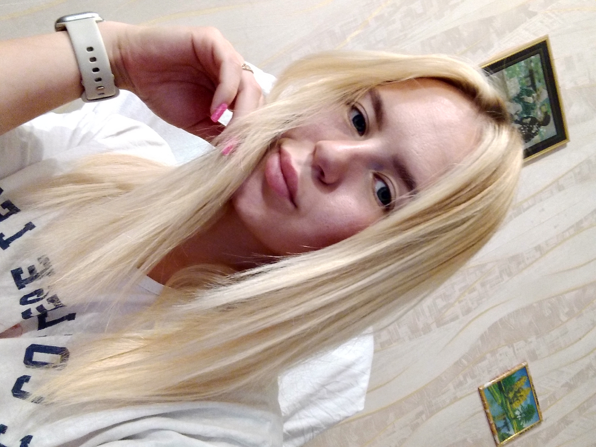 A young woman with long blonde hair is posing for a selfie in a room with patterned wallpaper. She is wearing a white t-shirt with dark lettering on it and a white smartwatch on her left wrist. Her right hand is resting on her chin. There are two small framed pictures on the wall behind her. The overall impression is a casual indoor setting.
