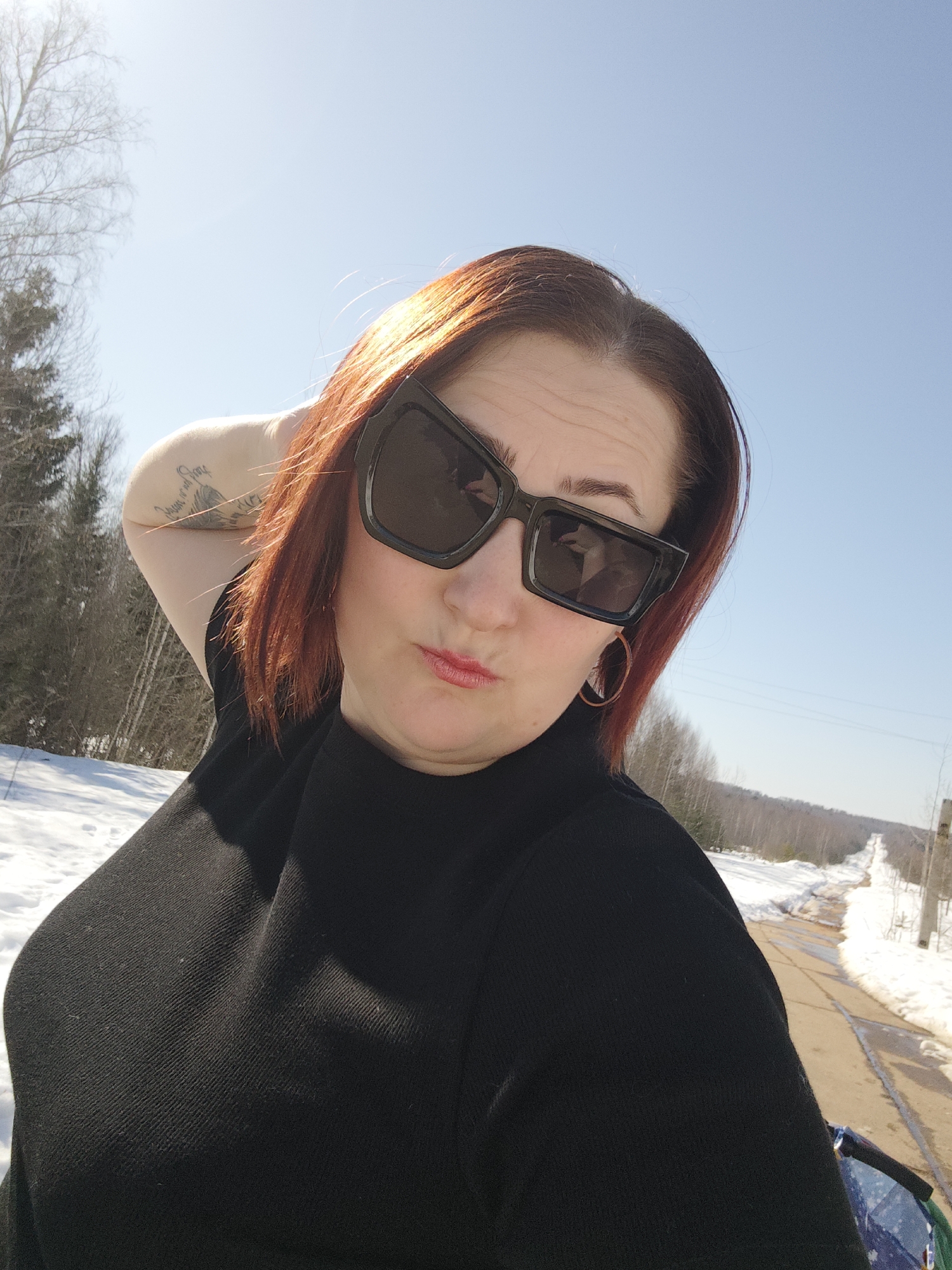 A woman with auburn hair and sunglasses is posing for a selfie on a sunny day. She wears a black top, and has her left arm raised with her hand behind her head. A tattoo is visible on her arm. The backdrop features a partly snowy road leading through a landscape of trees and melting snow. Based on the background, the location is likely Марковский, Russia.