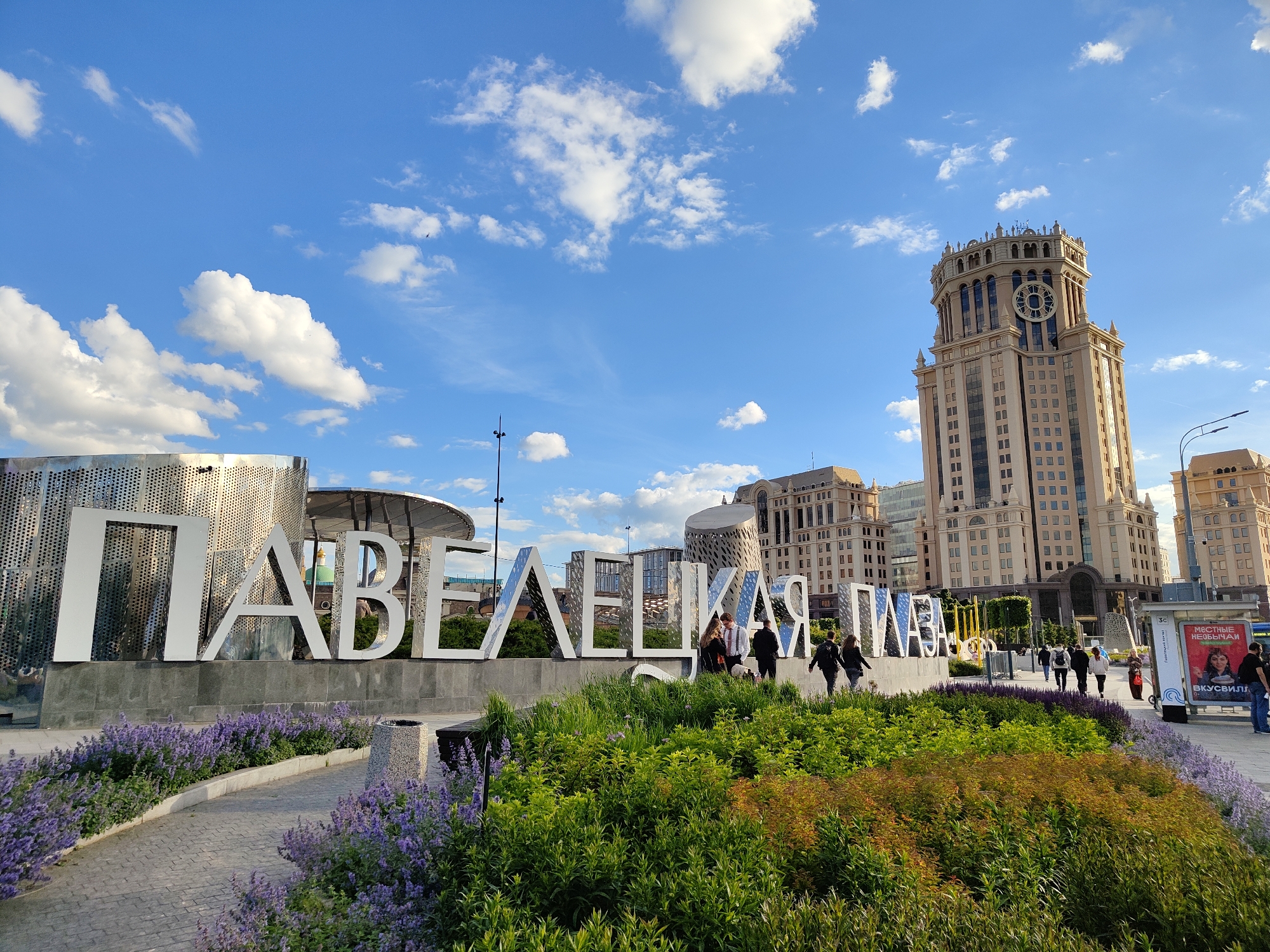 The image captures a street scene in Moscow, Russia, featuring the large white letters of the "PAVELETSKAYA" landmark. The letters are positioned in front of a garden bed filled with lush green plants and purple flowers.
In the background, various buildings rise into the blue sky dotted with fluffy white clouds. A prominent building with a clock tower stands tall, indicating an urban architectural landscape. People are walking along the paved sidewalk adjacent to the garden bed. A bus stop with advertisements is visible on the right side of the image.