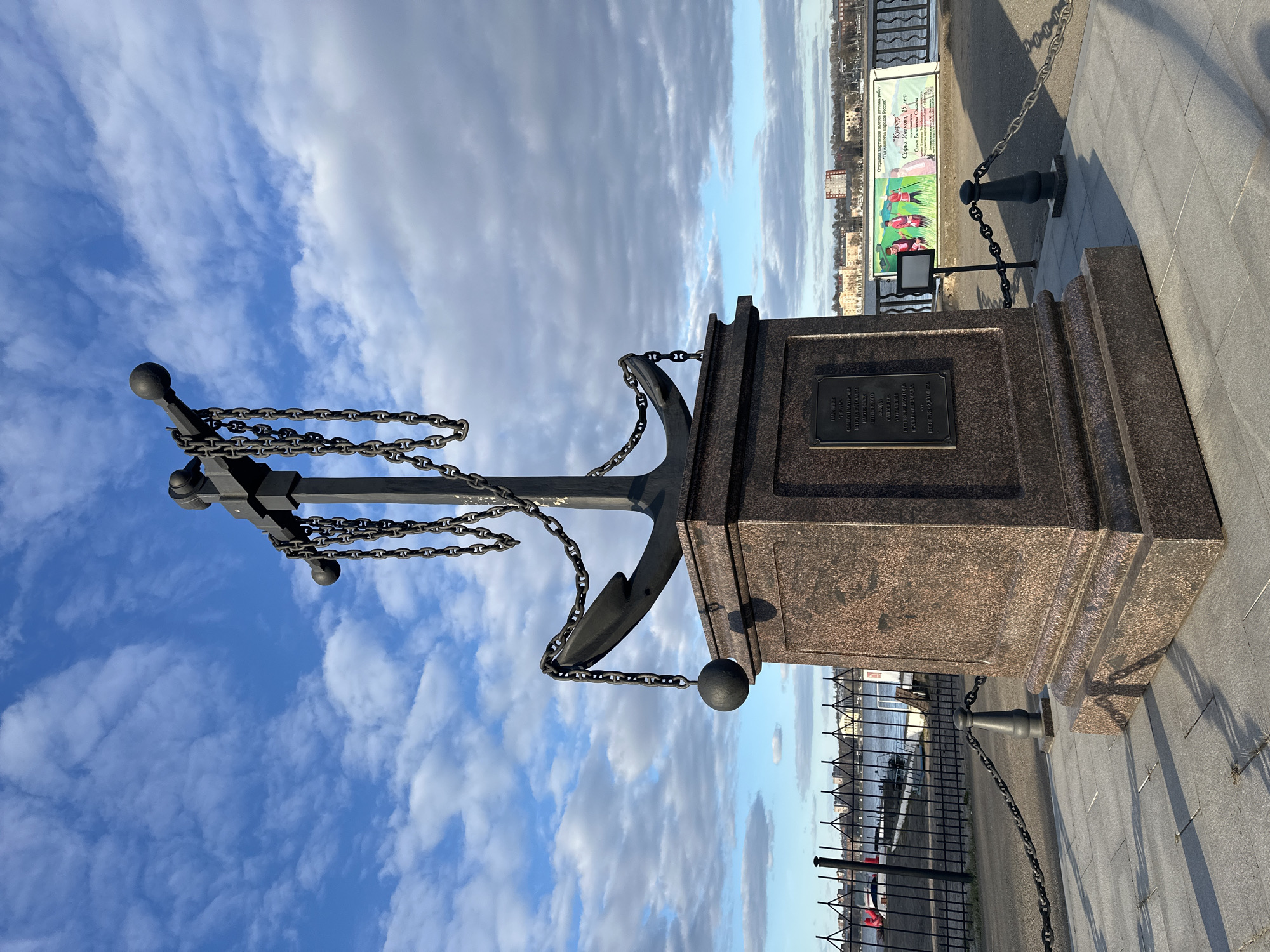 The image shows a monument in Votkinsk, Russia. The monument consists of an anchor on top of a granite base, surrounded by a chain-link fence. The anchor is black and is suspended above the base by chains. The granite base has a plaque on it with Cyrillic text. In the background, there is a building. A sign can be seen in the background for the "Kuarsur" drawing competition by Sofia Ivanova, 15 years old, and Olga Vasilievna Smalyanny. The sky is blue with some clouds. The monument is located on a paved area.