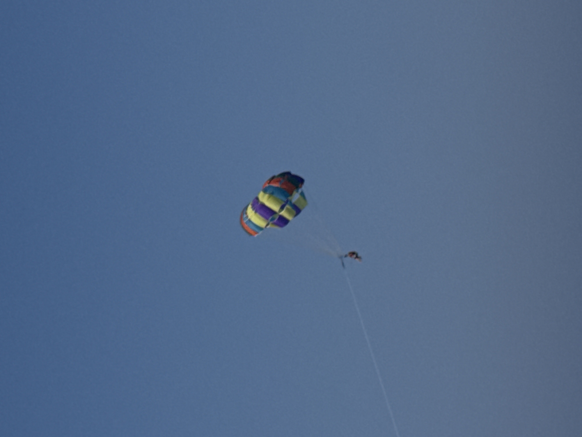 A colorful parasail, with sections of purple, yellow, and blue, is suspended in a clear blue sky. A person is attached to the parasail by a tow rope, which extends downwards and to the left. The person appears to be wearing a red or orange garment. The scene is likely Fethiye, Turkey, known for parasailing over its turquoise waters.