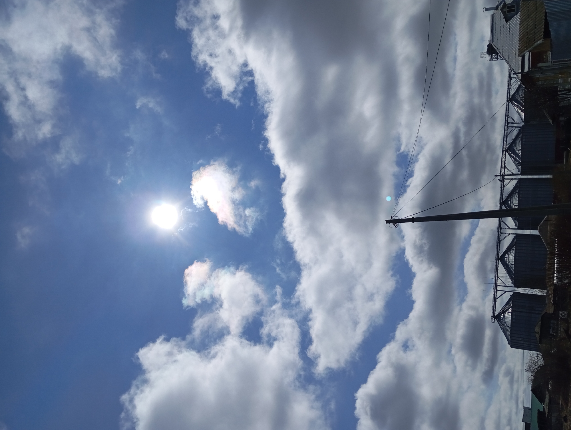 The image shows a sunny sky with scattered clouds in Yurgamysh, Russia. The sun is visible on the left, partially obscured by a cloud. An electrical pole and wires run vertically along the right side of the frame, adjacent to metal silos and buildings. The horizon is tilted slightly.