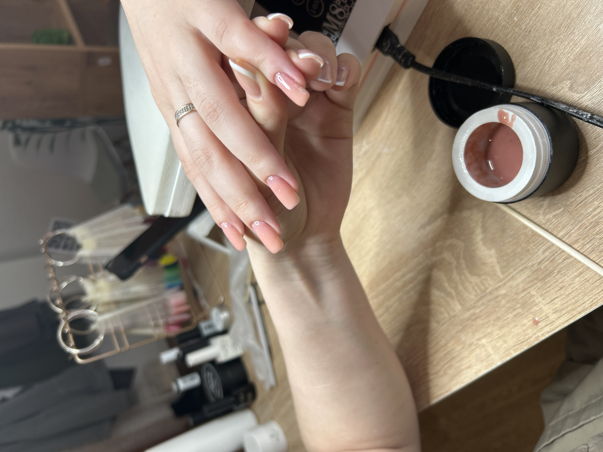 The image shows a close-up view of a person's hands with freshly manicured nails. The nails are square-shaped, with a nude or light pink base and a white tip on some nails. One of the hands has a ring on the ring finger. Adjacent to the hands is a jar of nail polish, presumably the one used for the manicure. To the side, there is a black power cord. There is a nail lamp, manicure tools, and other bottles of nail polish, all set against a light wooden surface that might be a table or desk. The setting suggests a nail salon, potentially in Chaikovsky, Russia. The primary subjects are the manicured hands and the associated manicure tools and materials.