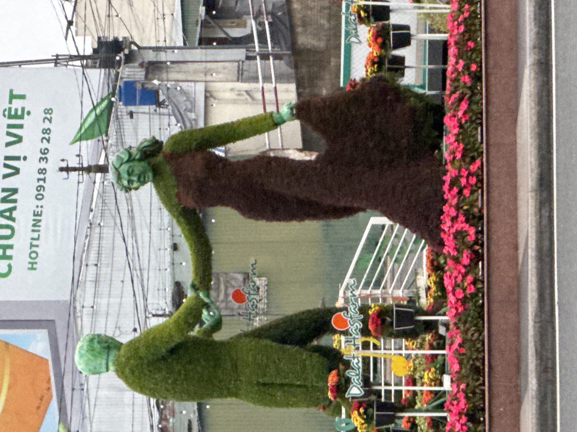 The image depicts a street scene in Da Lat, Vietnam, featuring two topiary sculptures of humans dancing. The sculptures are made of greenery, with one figure dressed in a green suit and the other in a brown, flowing dress. They are positioned in front of a building with a large sign that reads "HOTLINE: 0918 36 28 28". In the foreground, there is a flower bed with colorful blooms, and a white railing. The background shows more buildings and overhead wires. The overall impression is of an outdoor decorative display.
