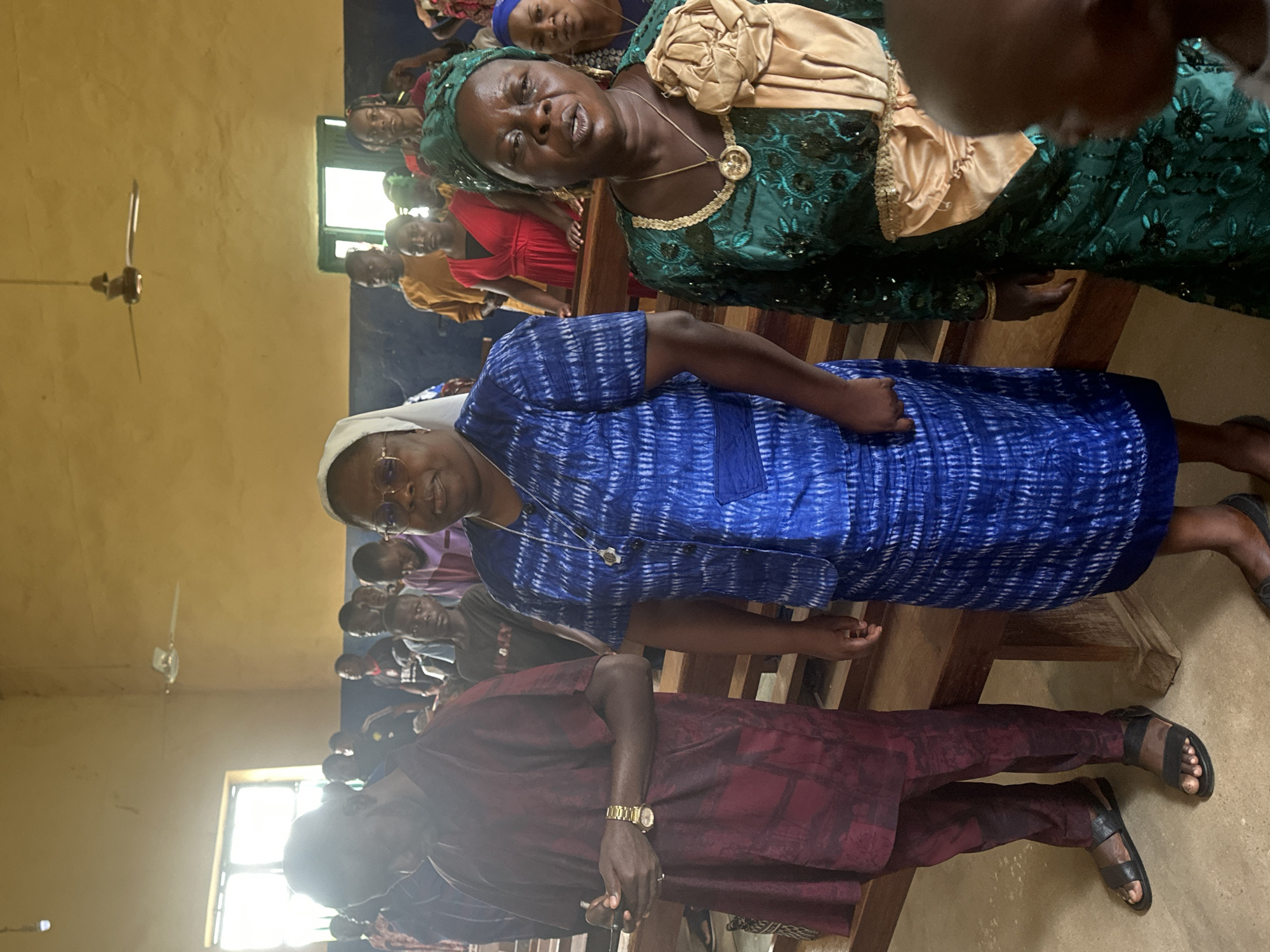 In a room with yellow walls, people are gathered. A ceiling fan is visible in the upper center of the image. The room appears to be a place of worship or gathering, possibly a church hall in Akwanga, Nigeria. The lighting suggests it is daytime, with natural light coming from windows on the left side.
In the foreground, a man on the left wears a maroon, patterned robe and black sandals. He is looking down, with his hands clasped. To his right, a woman wearing glasses and a white head covering stands in a blue, patterned dress and skirt. She is looking forward. Further to the right, a woman in a green, sequined dress and headscarf faces the viewer, with an open mouth as if speaking or singing. She is adorned with a gold pendant necklace and a large gold fabric embellishment on her dress. Behind these individuals, other people, including children and adults, are visible standing and seated on wooden benches. The overall impression is of a communal gathering, possibly a service or event.