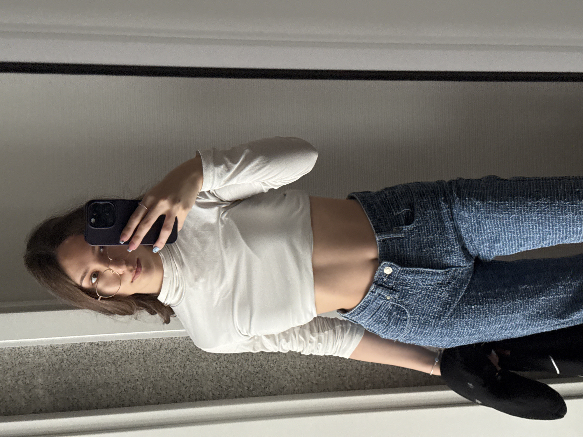 A young woman is taking a mirror selfie. She is wearing a white long-sleeved turtleneck crop top, blue tweed-like pants, and black shoes. She has brown hair and is wearing glasses. She is holding a dark-colored smartphone. The background appears to be a neutral-colored wall and a gray floor. The location cannot be determined from the image.