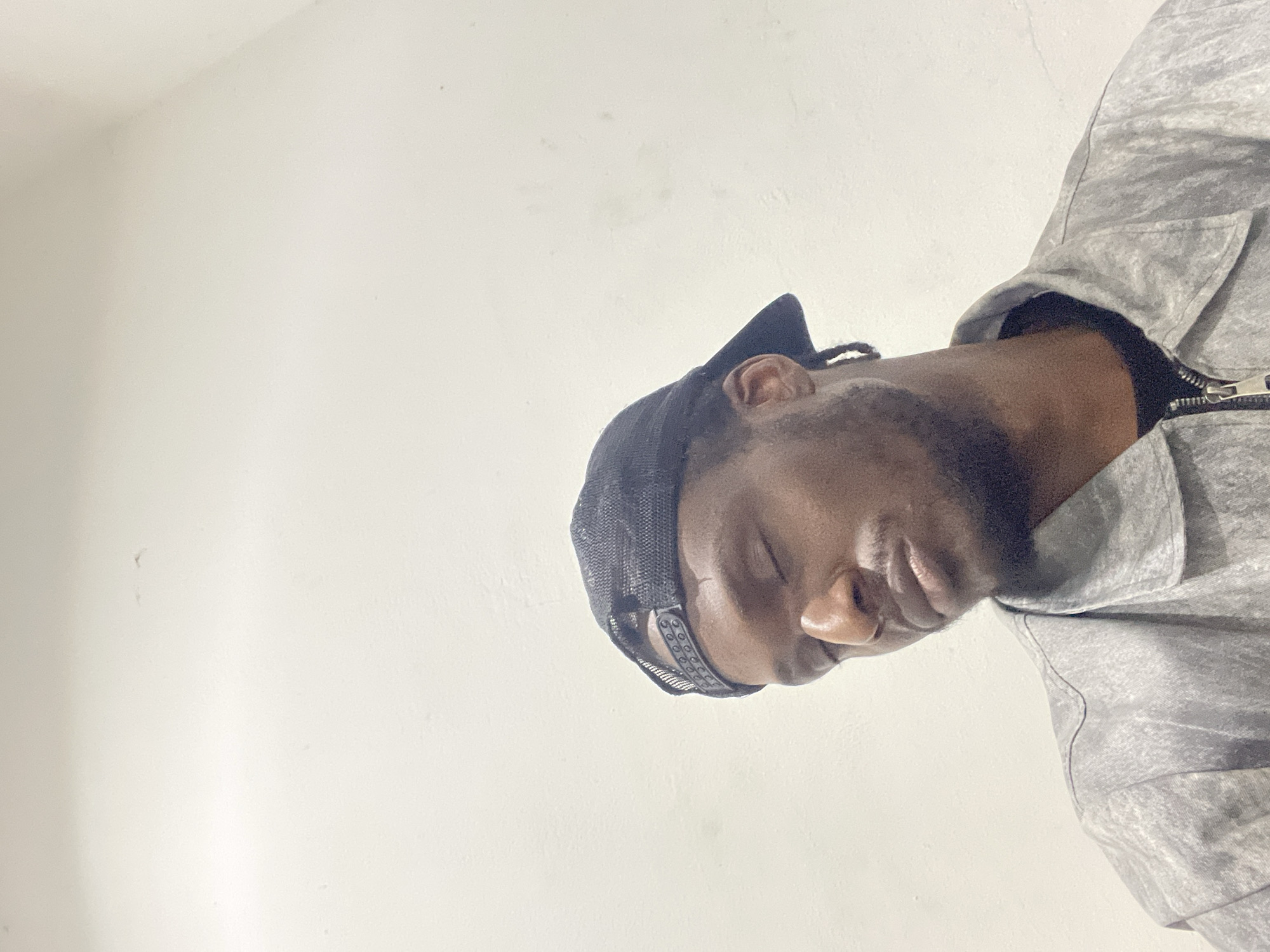 A close-up, low-angle shot shows a young Black man with his eyes closed, wearing a black baseball cap and a grey zip-up jacket over a black t-shirt. The man has a slight beard and mustache. The background is a plain, off-white wall. The lighting appears to be artificial, casting soft shadows on the man's face and the wall. There are no discernible landmarks or architectural features to indicate a specific location in Abuja, Nigeria. The overall impression is that of a candid, perhaps spontaneous, portrait.